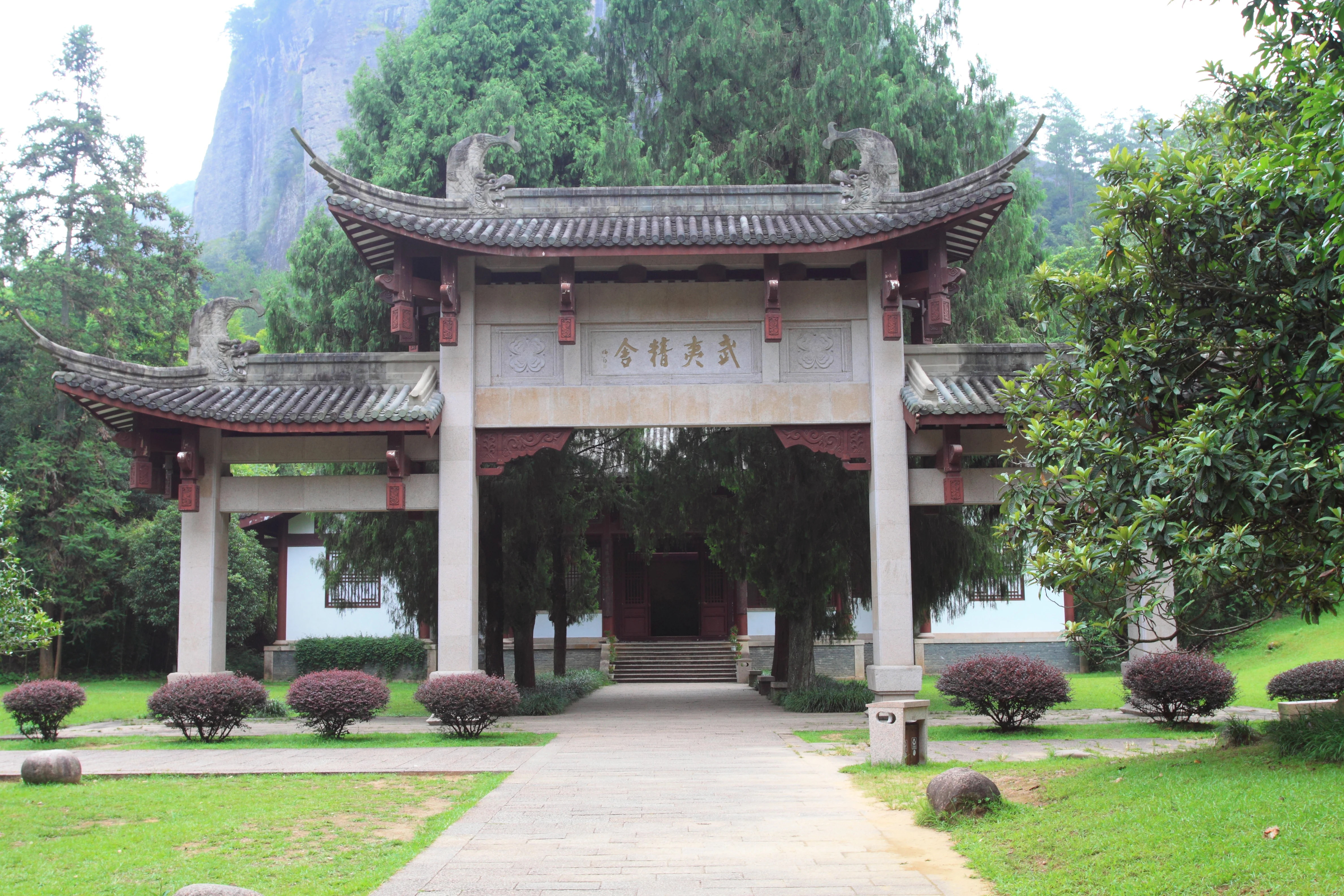 A traditional Chinese arched gateway with tiled, upturned eaves, set in a garden with lawn and trees, framing a path leading to a building.