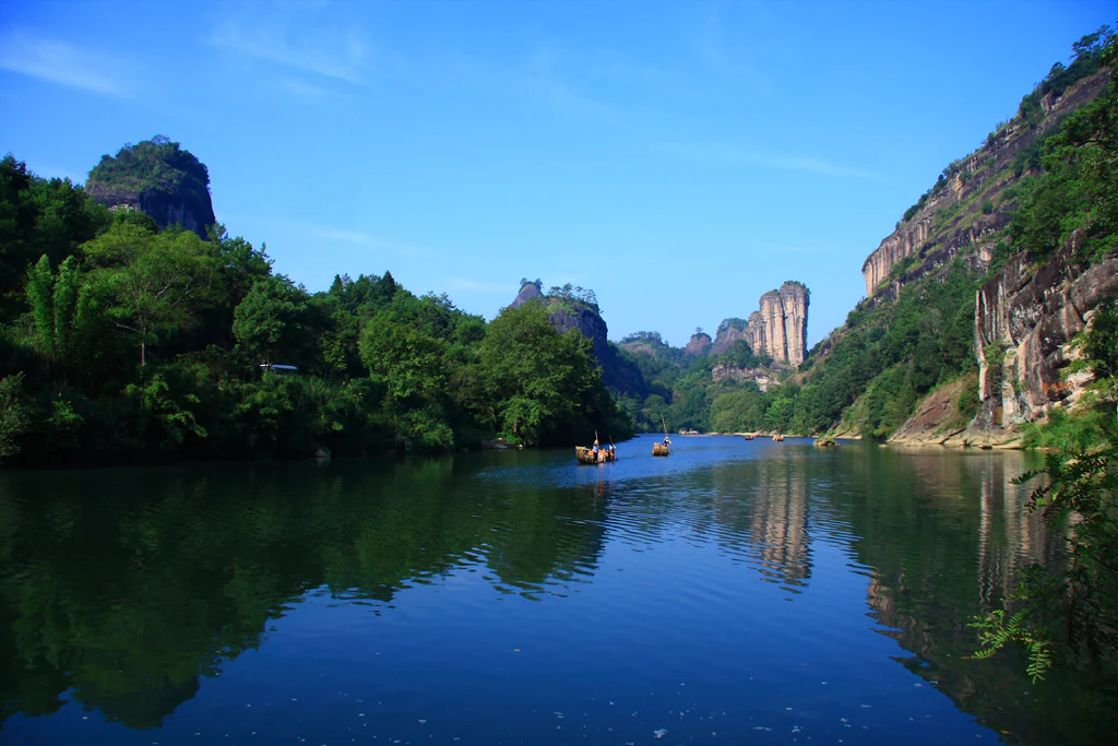 A wide view of a calm, reflective river flowing between steep, lush, rocky mountains in Wuyishan, with small wooden boats carrying passengers in the distance.