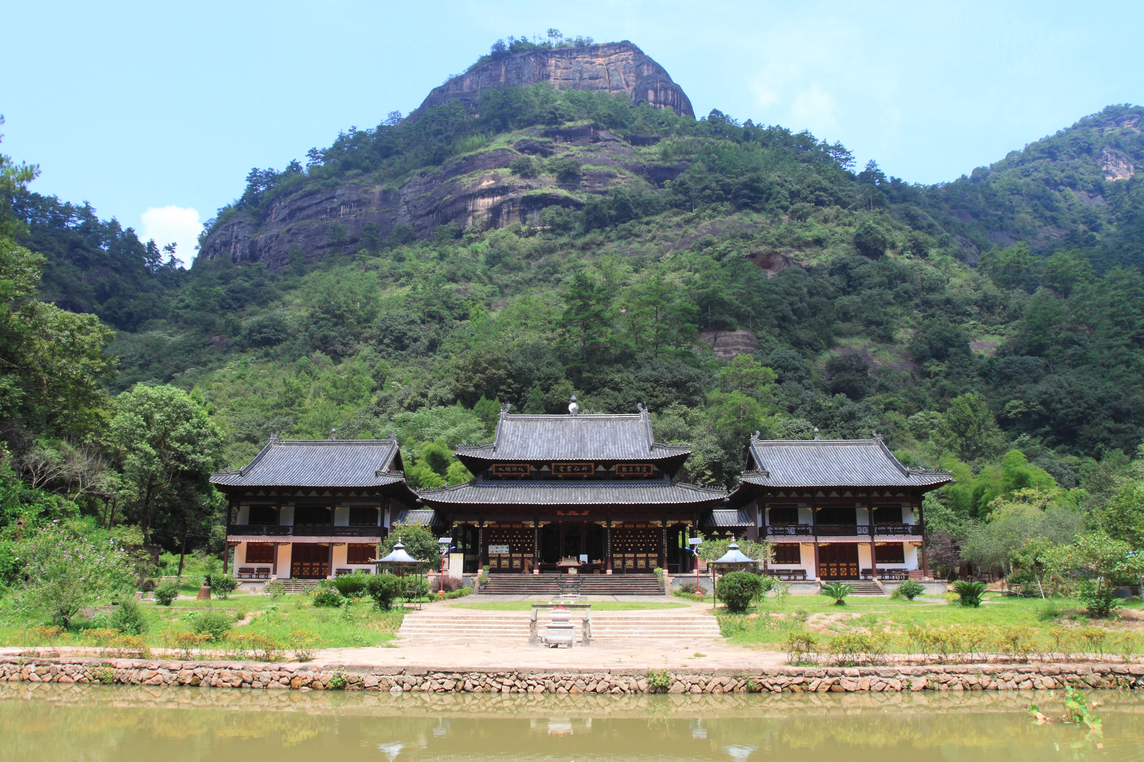 A traditional Chinese temple complex sits in front of a steep, tree-covered rocky mountain cliff, with a pond in the foreground.