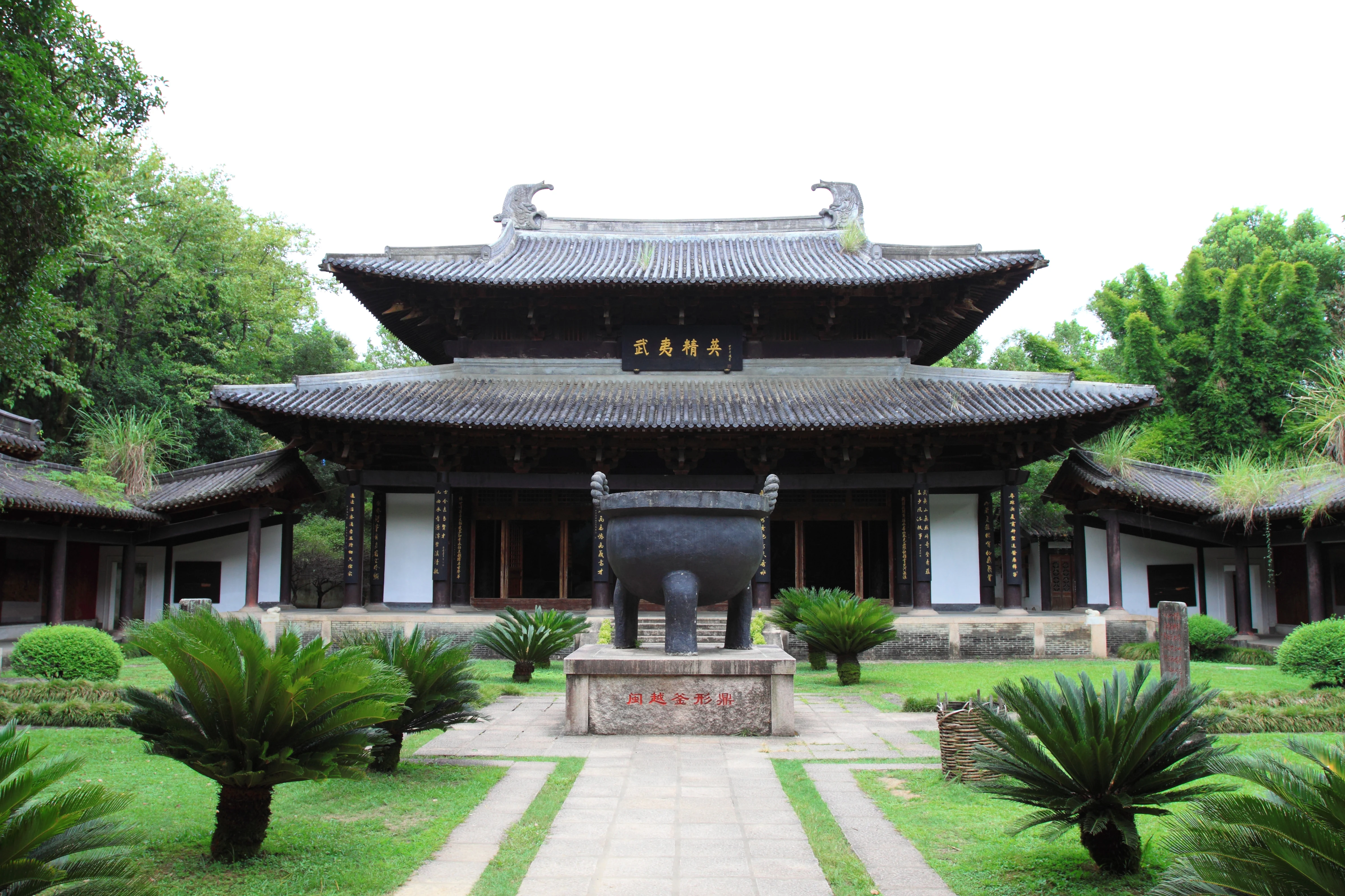 A traditional Chinese wooden building with dark tiled roofs, a central courtyard, and a large bronze tripod vessel sitting on a stone pedestal in the foreground, surrounded by cycad plants and lawn.