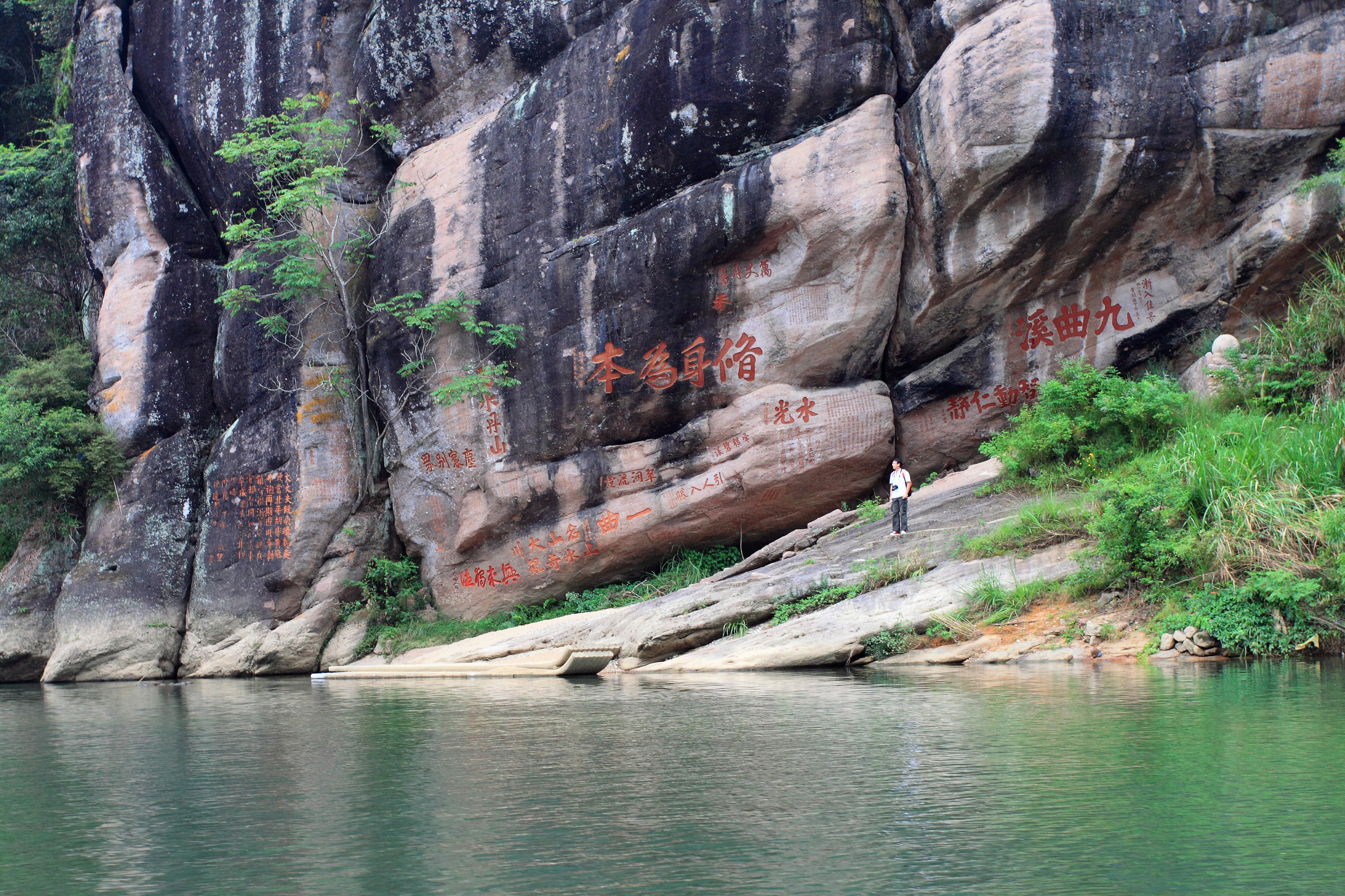 A wide view of a calm green river flowing past a large, dark-colored cliffside featuring reddish Chinese calligraphy carved directly into the rock face.