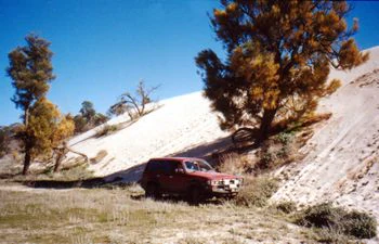 Red SUV on dirt road with sandy hill, mallee trees, and clear blue sky