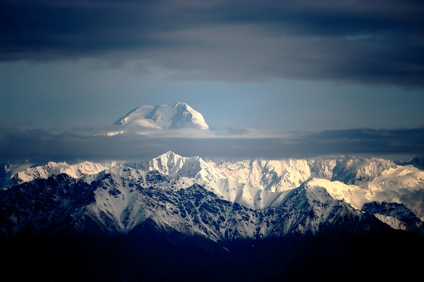 A dramatic, long-distance view of the snow-capped Tomur Peak rising above a secondary ridgeline of the Tianshan mountains under a dark, overcast sky.