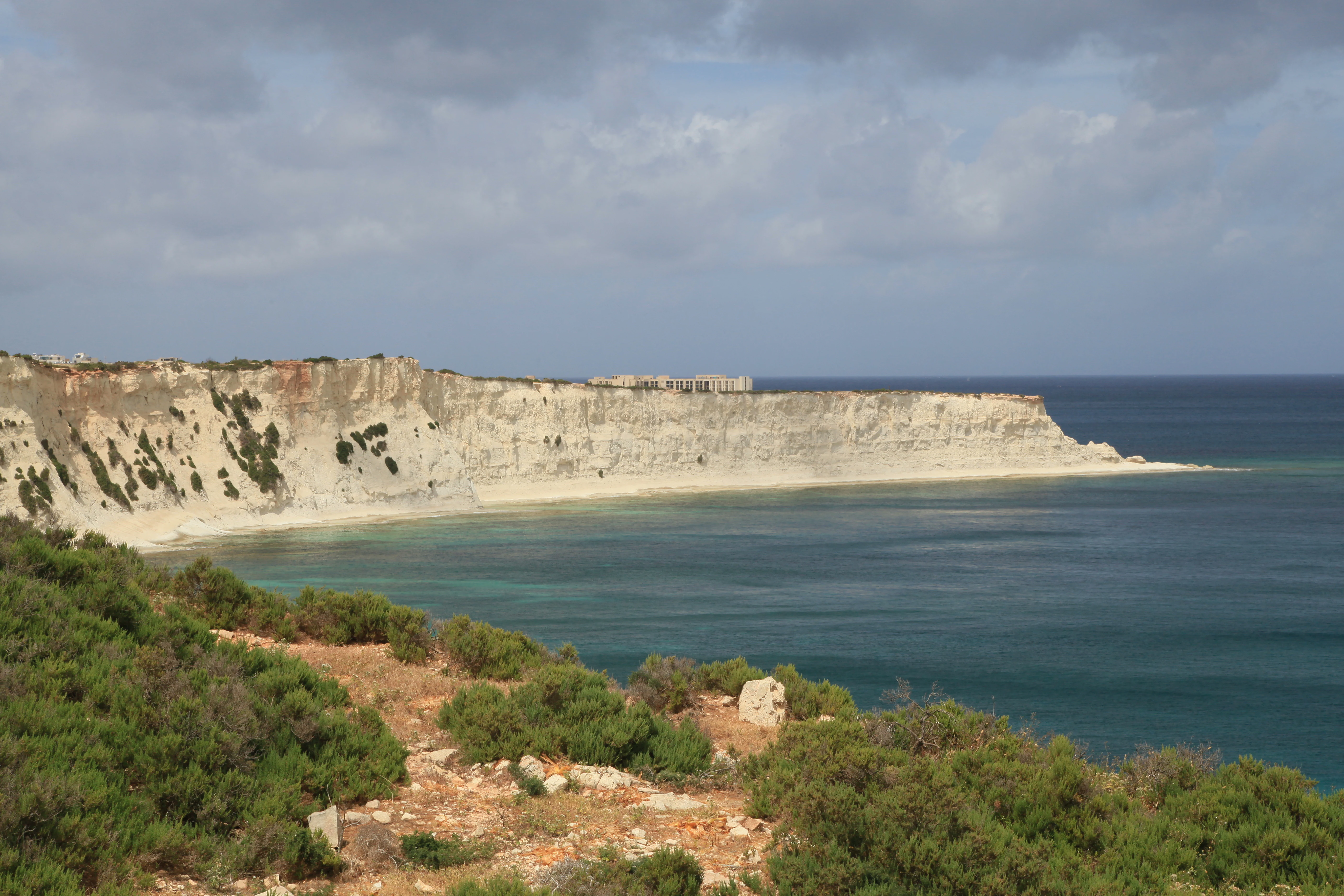 A wide coastal landscape showing limestone cliffs meeting the sea, with green Mediterranean scrubland in the foreground.