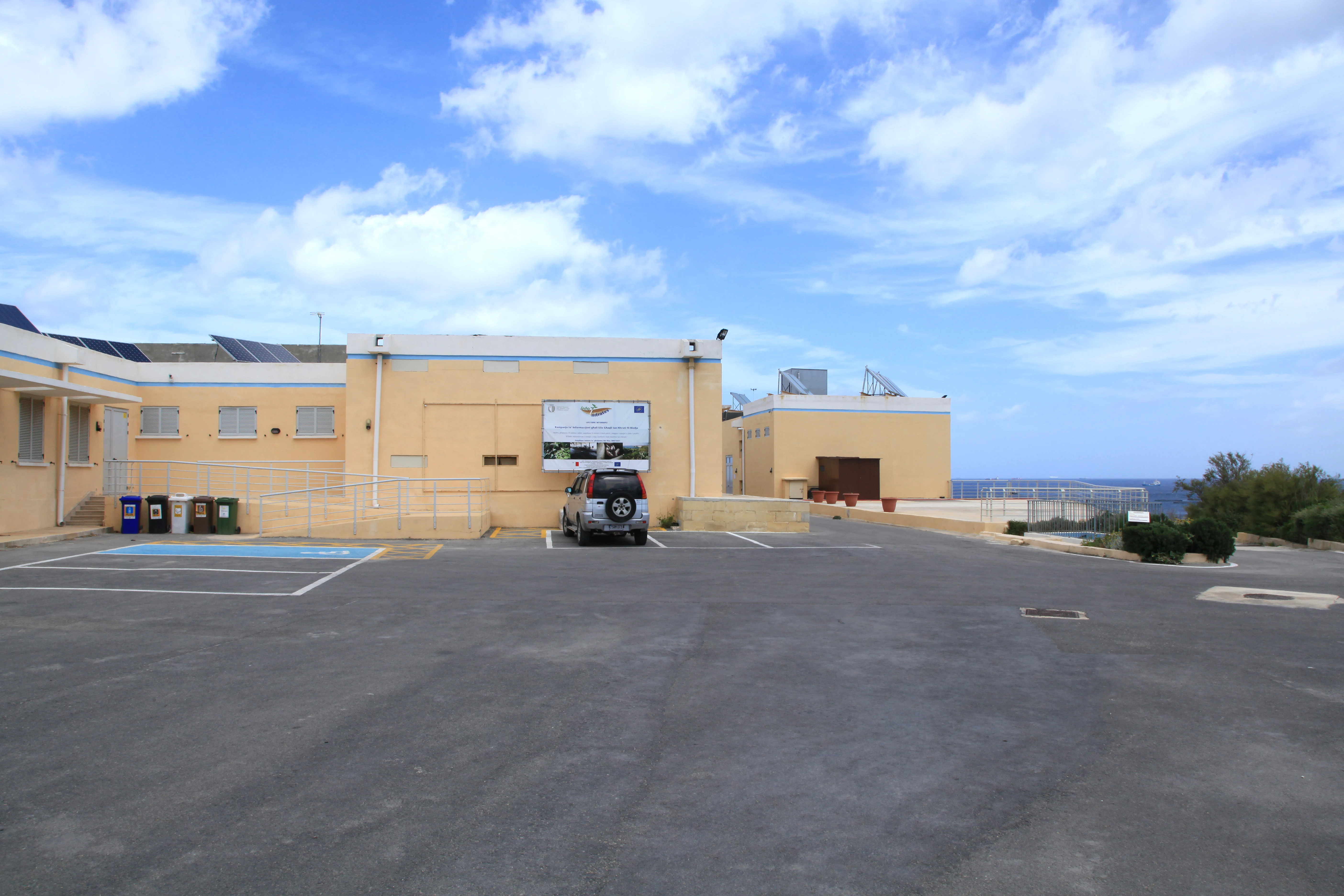 A wide outdoor view of a low, peach-colored building with a parking lot in front under a partly cloudy blue sky.