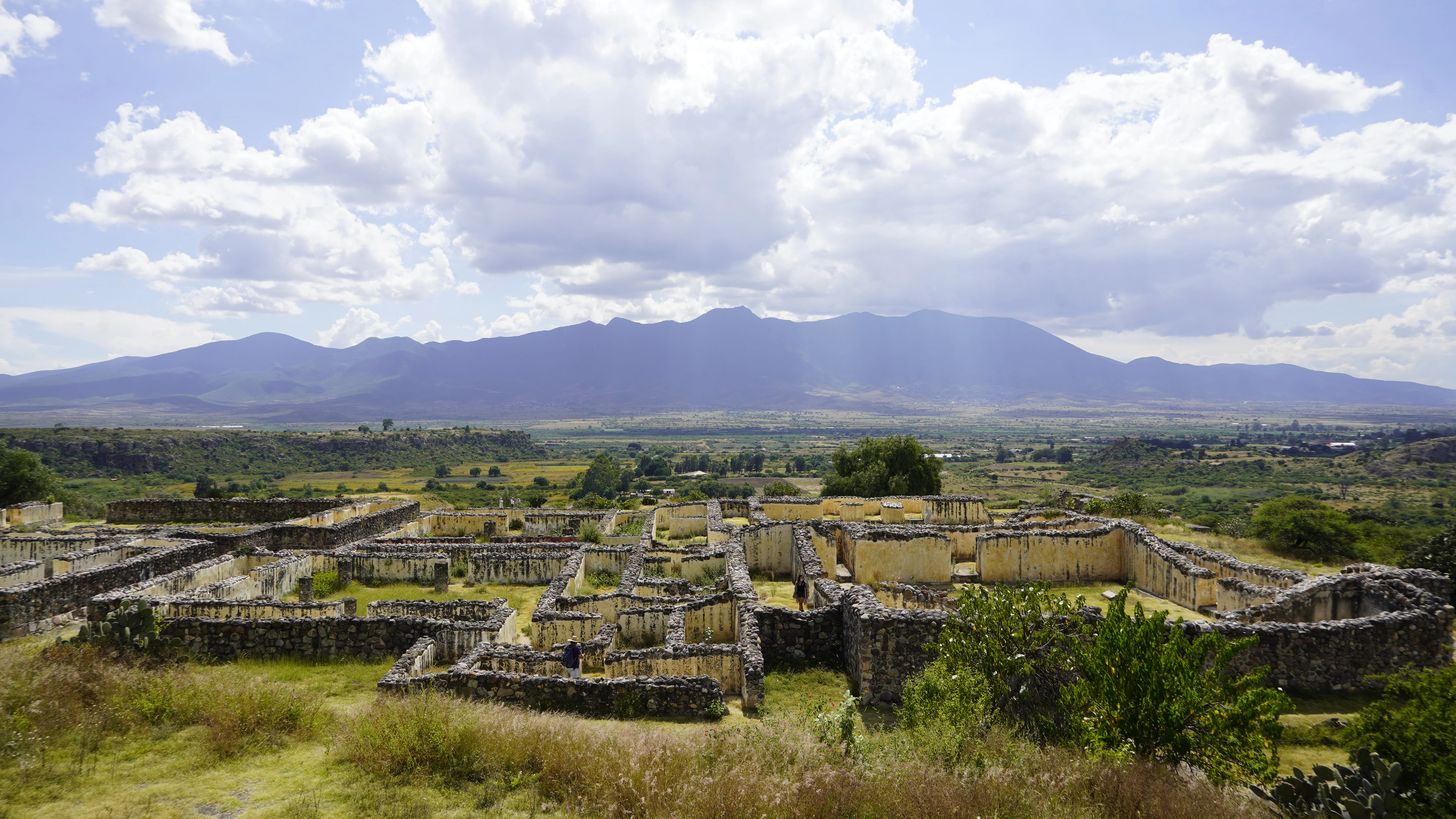 Una vista panorámica de gran angular de ruinas de muros de piedra zapotecas antiguas y cimientos de edificios en primer plano, con un valle extenso y grandes montañas al fondo bajo un cielo azul con nubes dispersas.