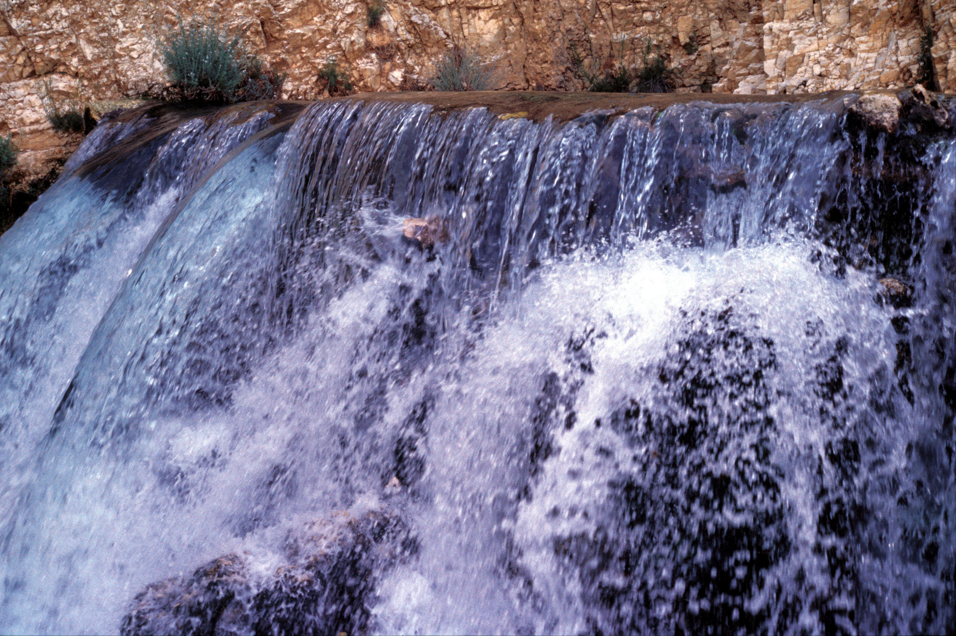 Una vista de cerca de una pequeña cascada que fluye sobre rocas, con un muro de piedra visible al fondo.