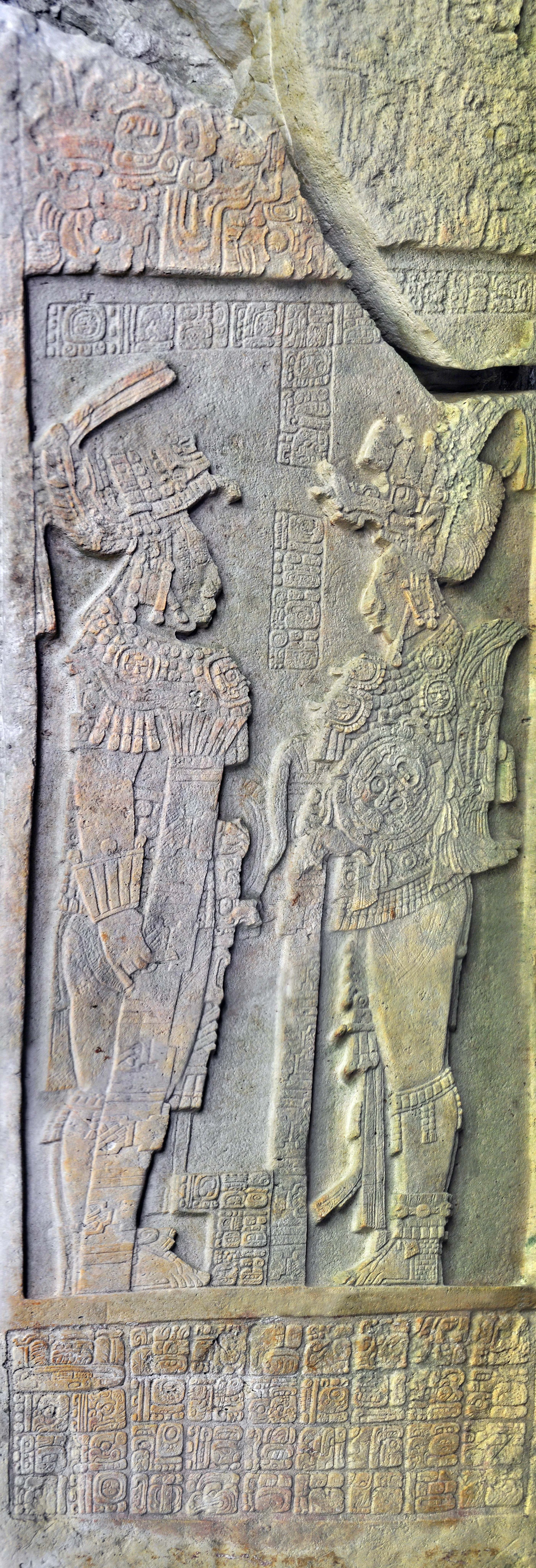 Ancient stone stela with intricate Mayan carvings of two figures in ceremonial attire surrounded by hieroglyphic inscriptions.