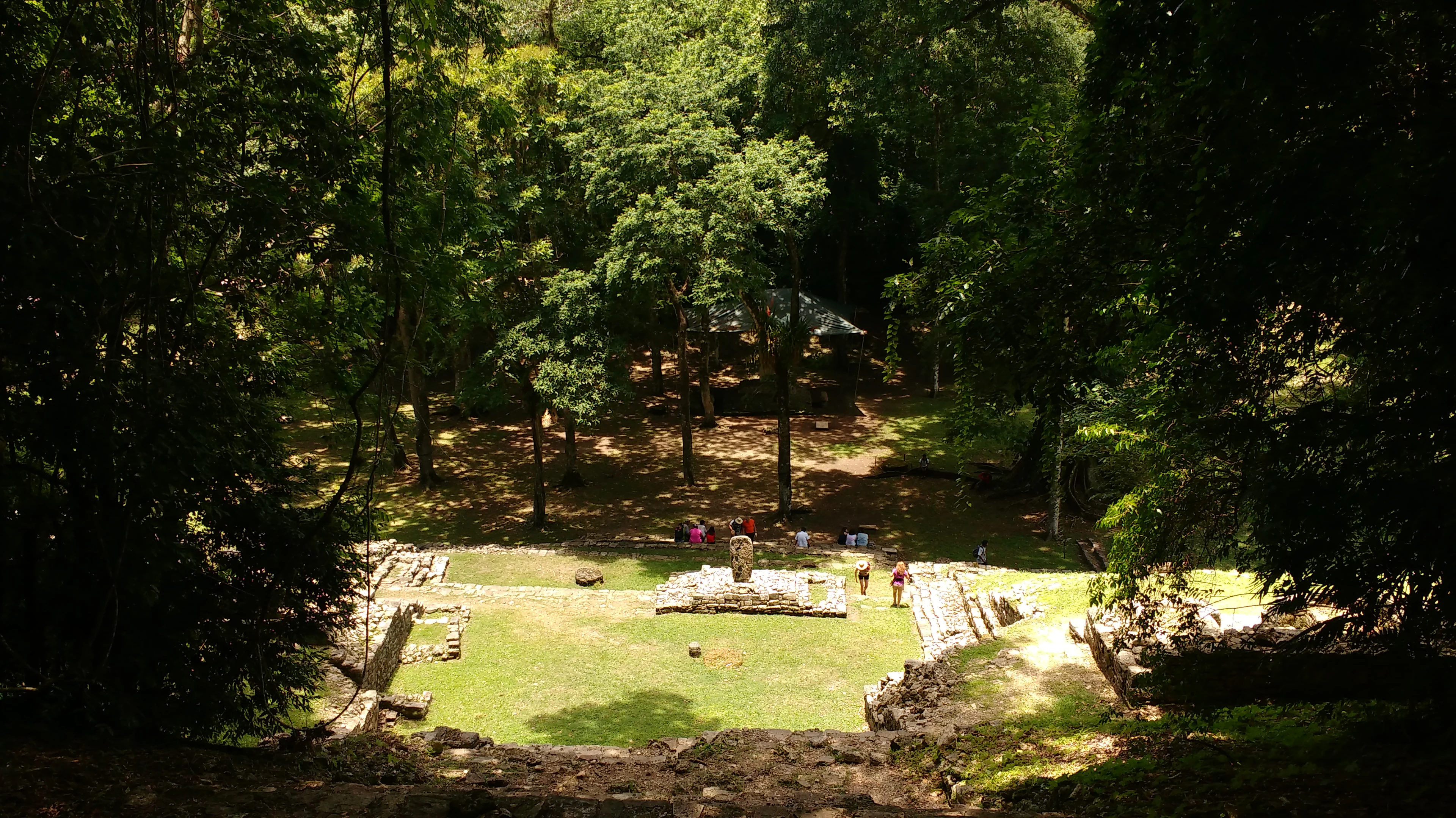 High-angle view looking down into a small grassy plaza surrounded by ancient stone ruins and thick tropical forest.