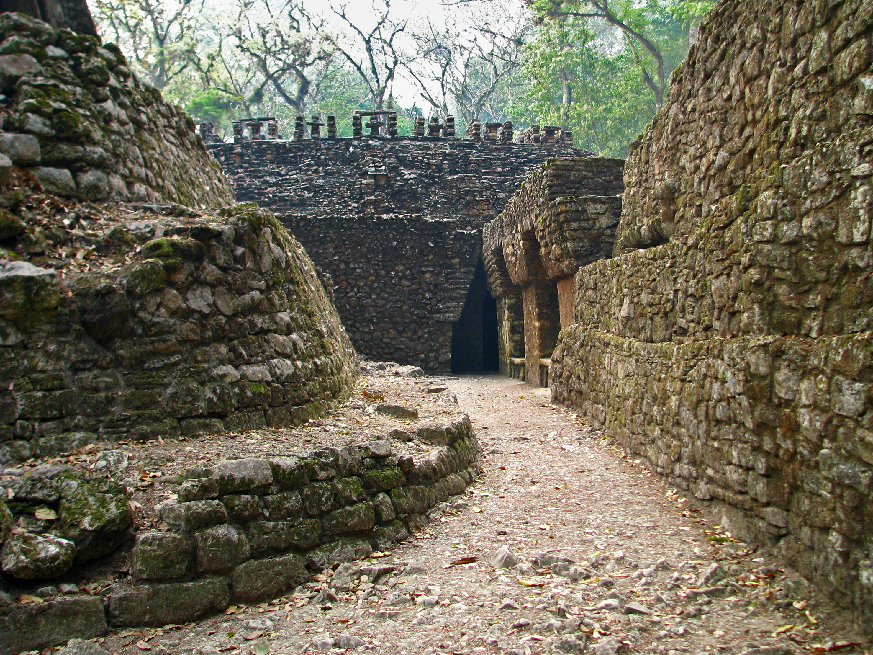 A path winds between ancient stone Maya ruins with a dark arched entryway visible in the distance at Yaxchilán.