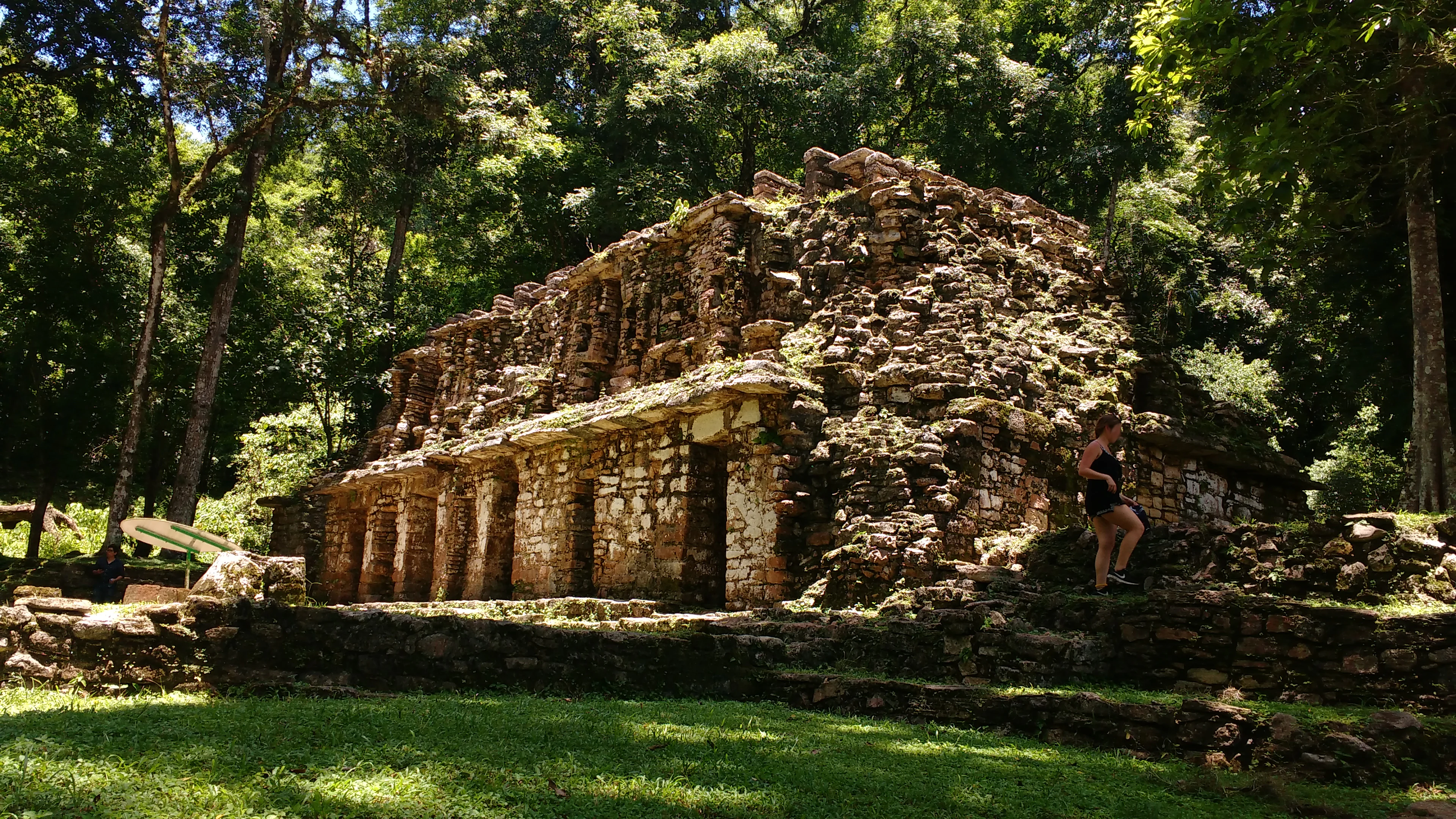 A weathered stone Maya structure with multiple doorways partially shaded by dense green jungle canopy, with a person standing on the right side.
