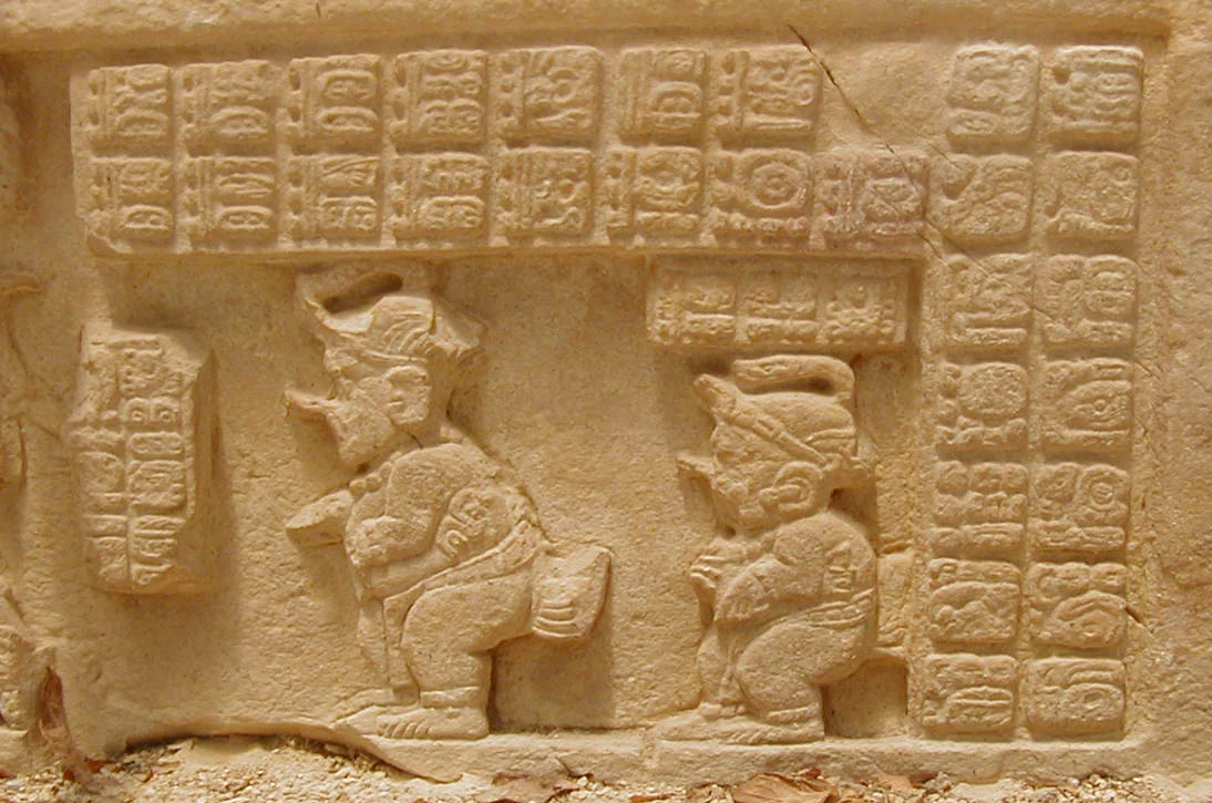A close up view of an ancient stone lintel carving at Yaxchilan showing two small figures, possibly dwarfs, depicted in profile alongside blocks of carved Maya hieroglyphs.