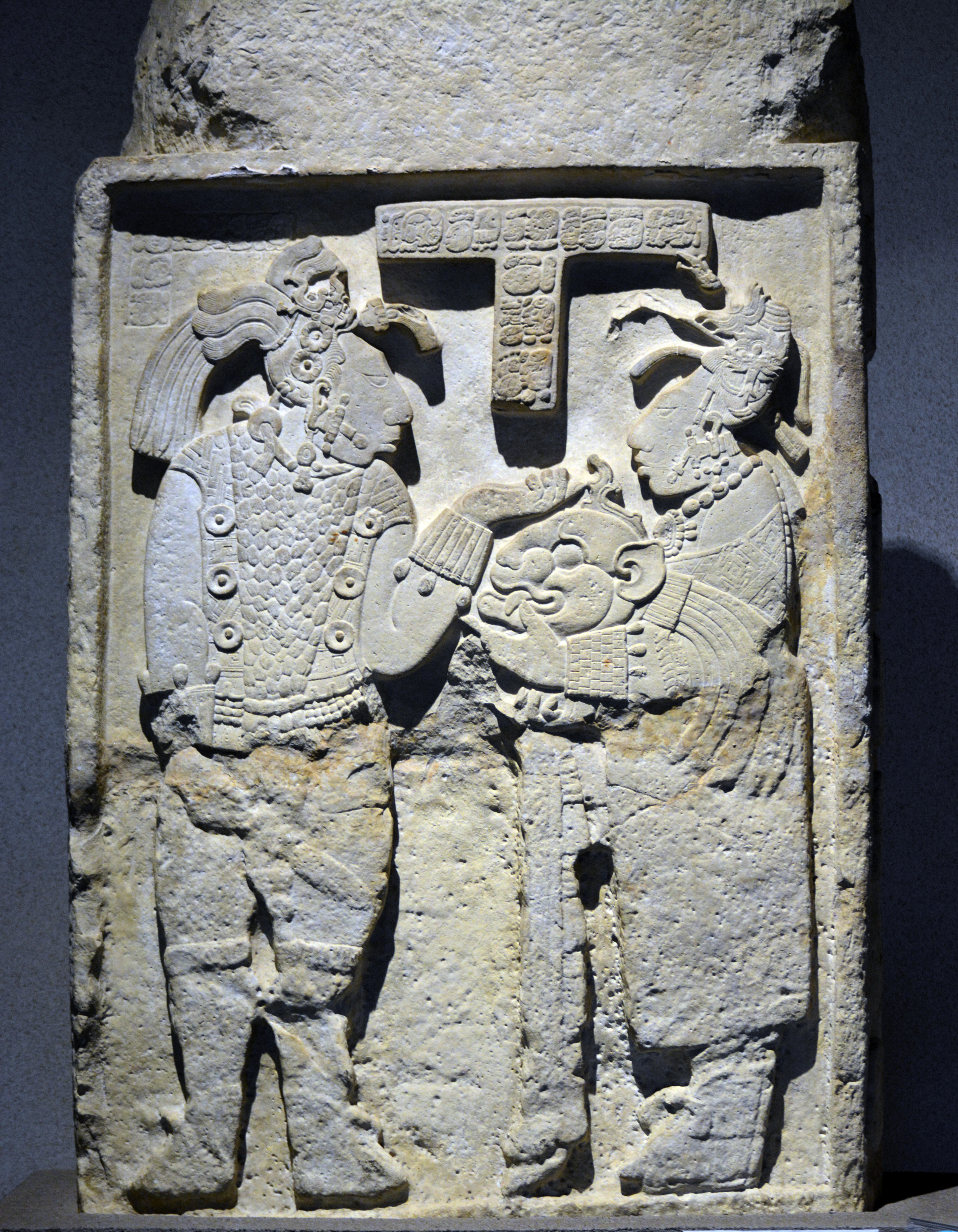 A vertically oriented rectangular limestone lintel featuring detailed relief carvings of two human figures in profile, including a ruler and consort, with Maya hieroglyphs at the top center.