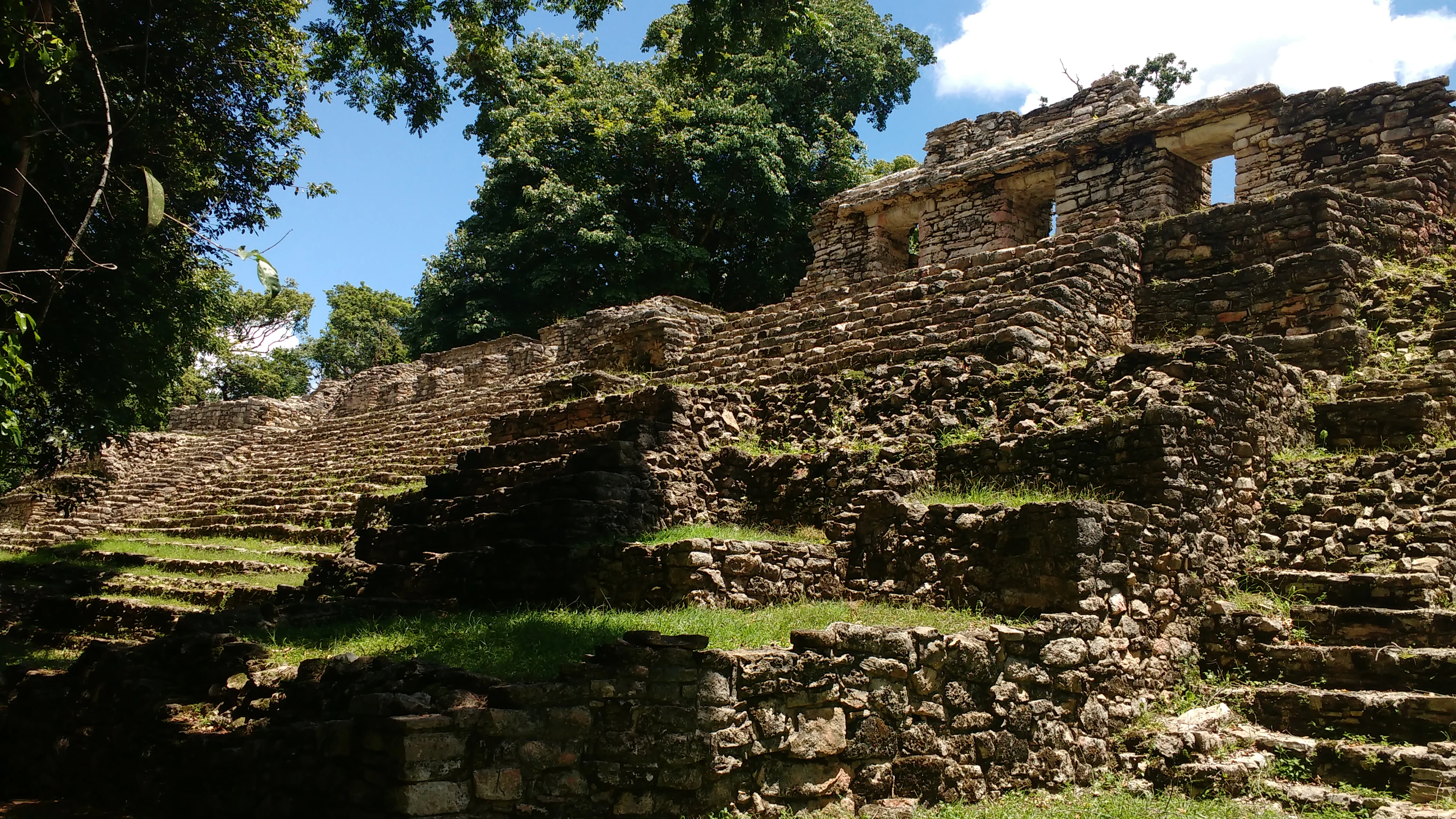 A low-angle view of weathered stone ruins featuring a terraced pyramid base and arched stone doorways, surrounded by trees under a blue sky.