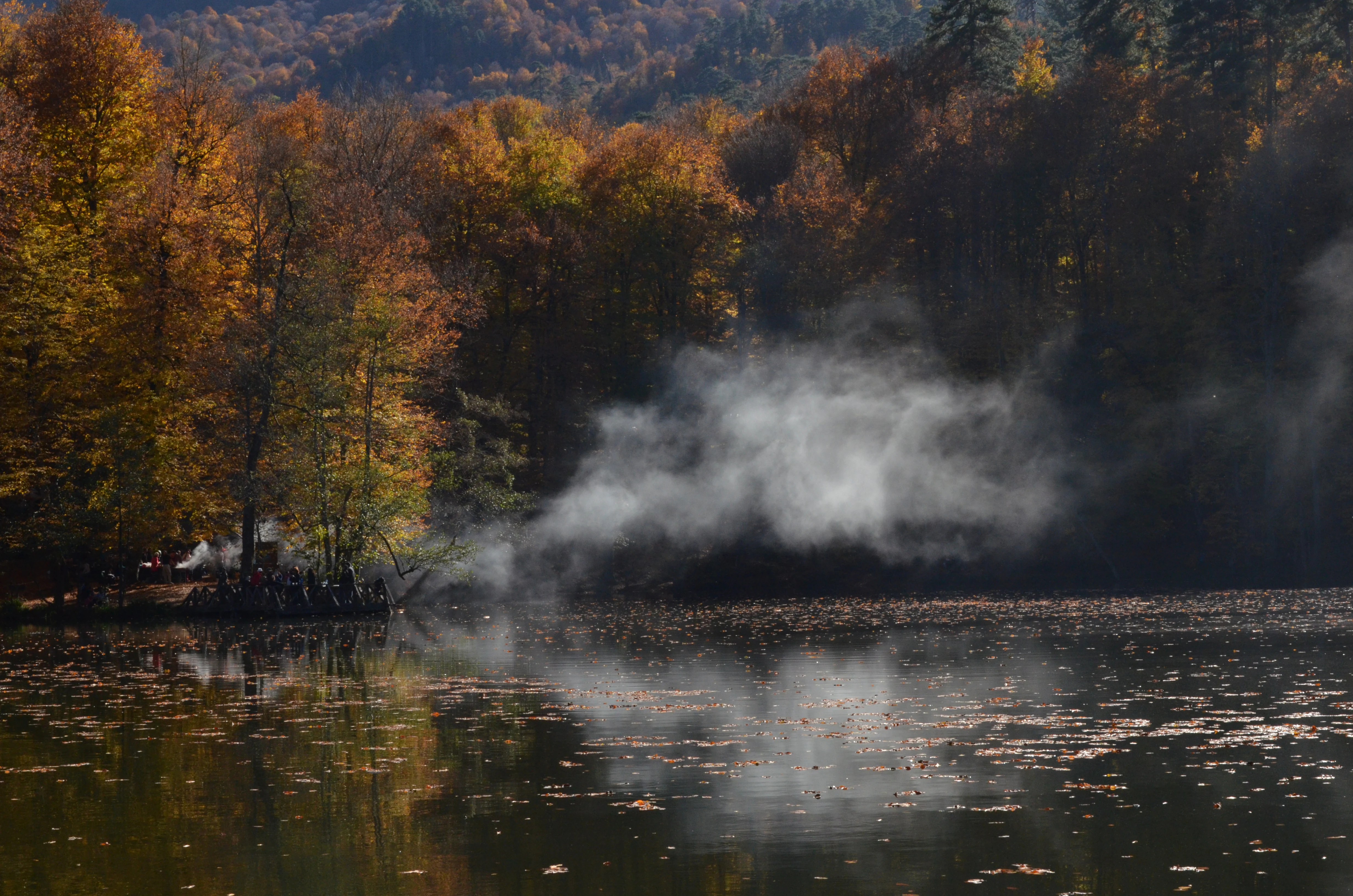 A lake with a wooden pier extending over calm water, surrounded by trees with orange and yellow foliage, and mist rising from the water surface