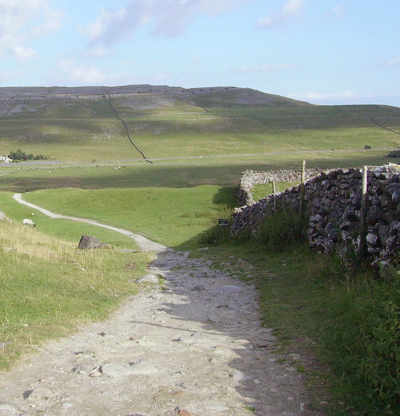 Dry stone wall with gravel path, grassy fields, and hills in background