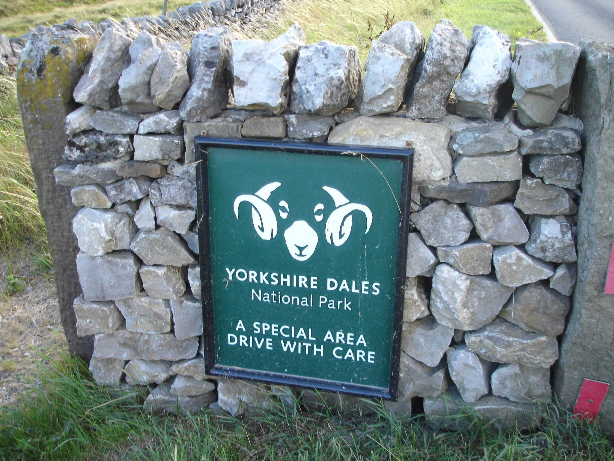 Green sign with white text and sheep logo reading 'Yorkshire Dales National Park A SPECIAL AREA DRIVE WITH CARE' mounted on a stone wall with grass in the background