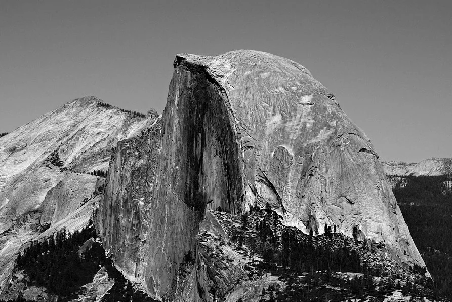 Black and white photograph of Half Dome rock formation with steep cliffs and forested slopes under clear sky.