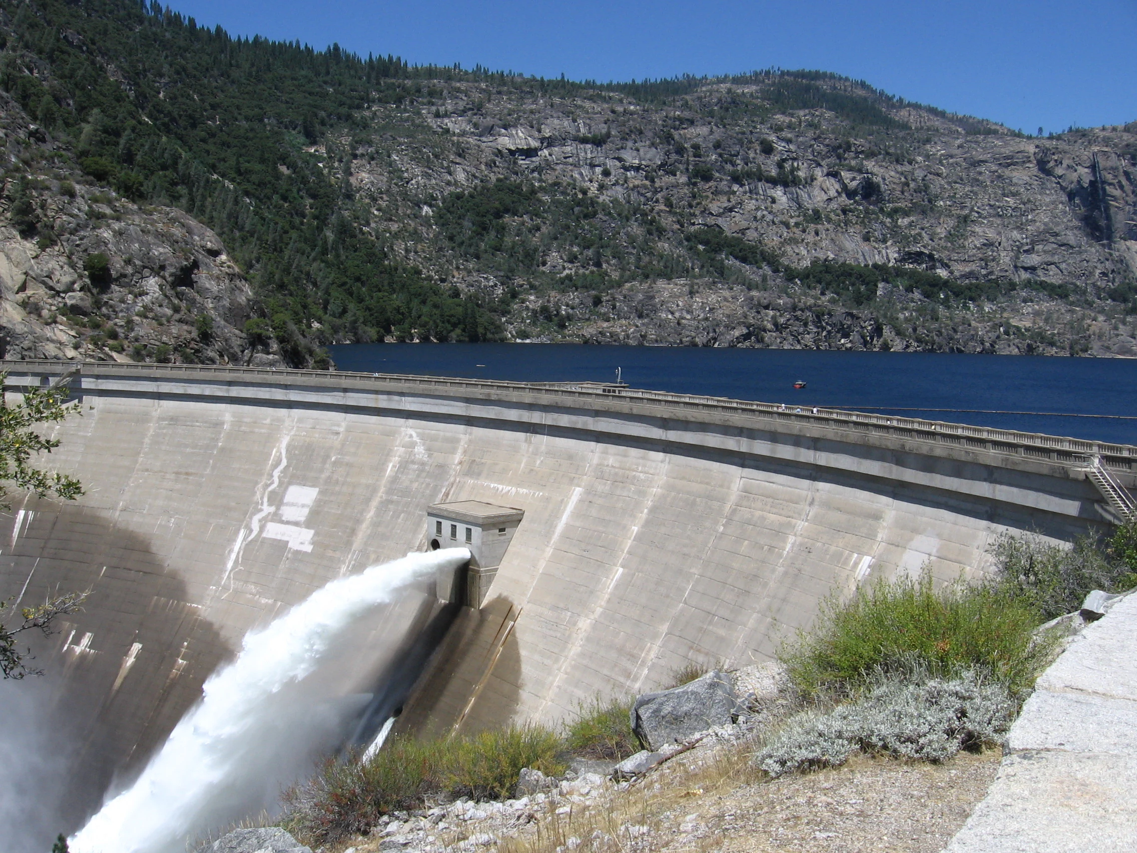 Concrete dam with water cascading from spillway, surrounded by rocky hills and a lake under clear blue sky.