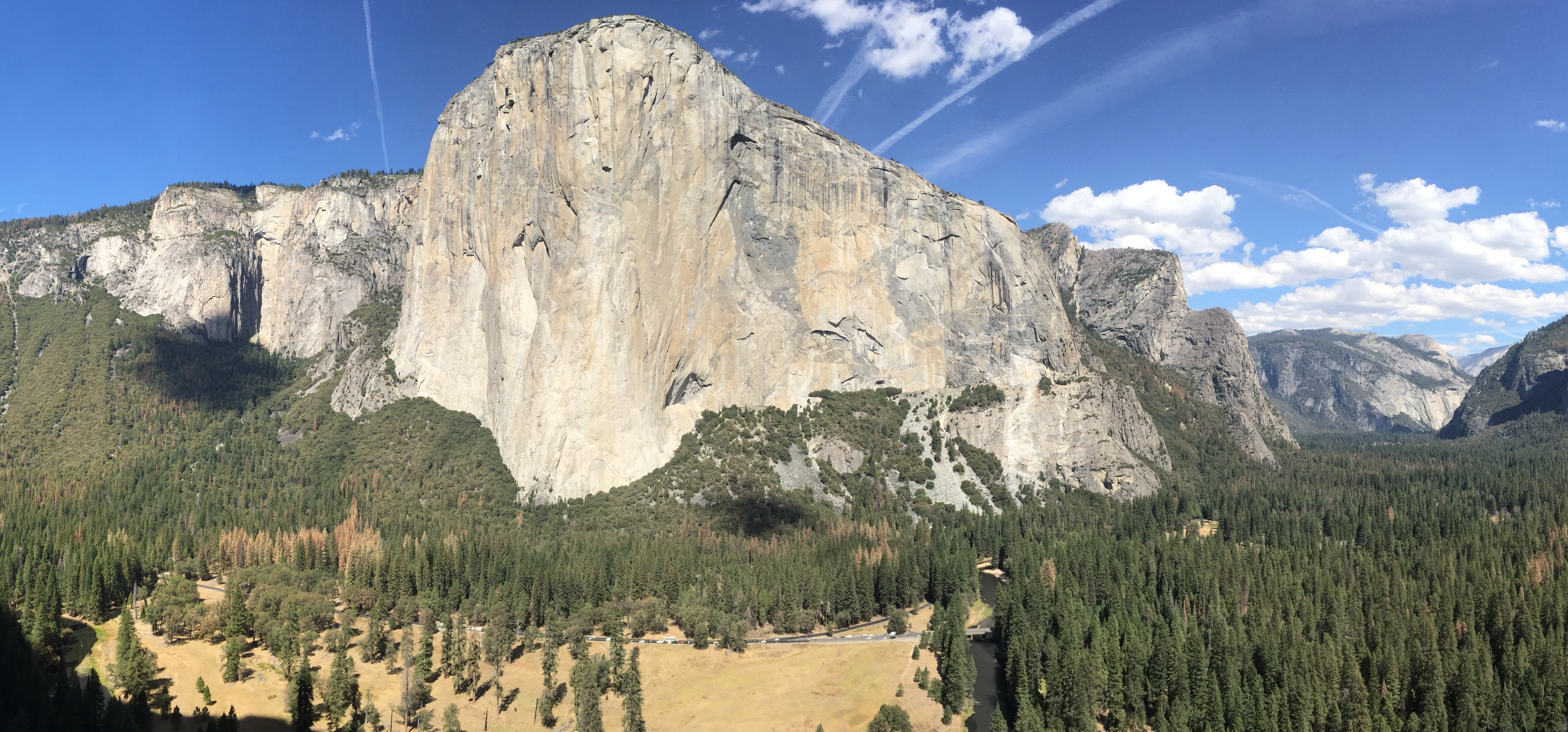 Wide panoramic view of El Capitan rock formation in Yosemite Valley with forested hills and clear blue sky.