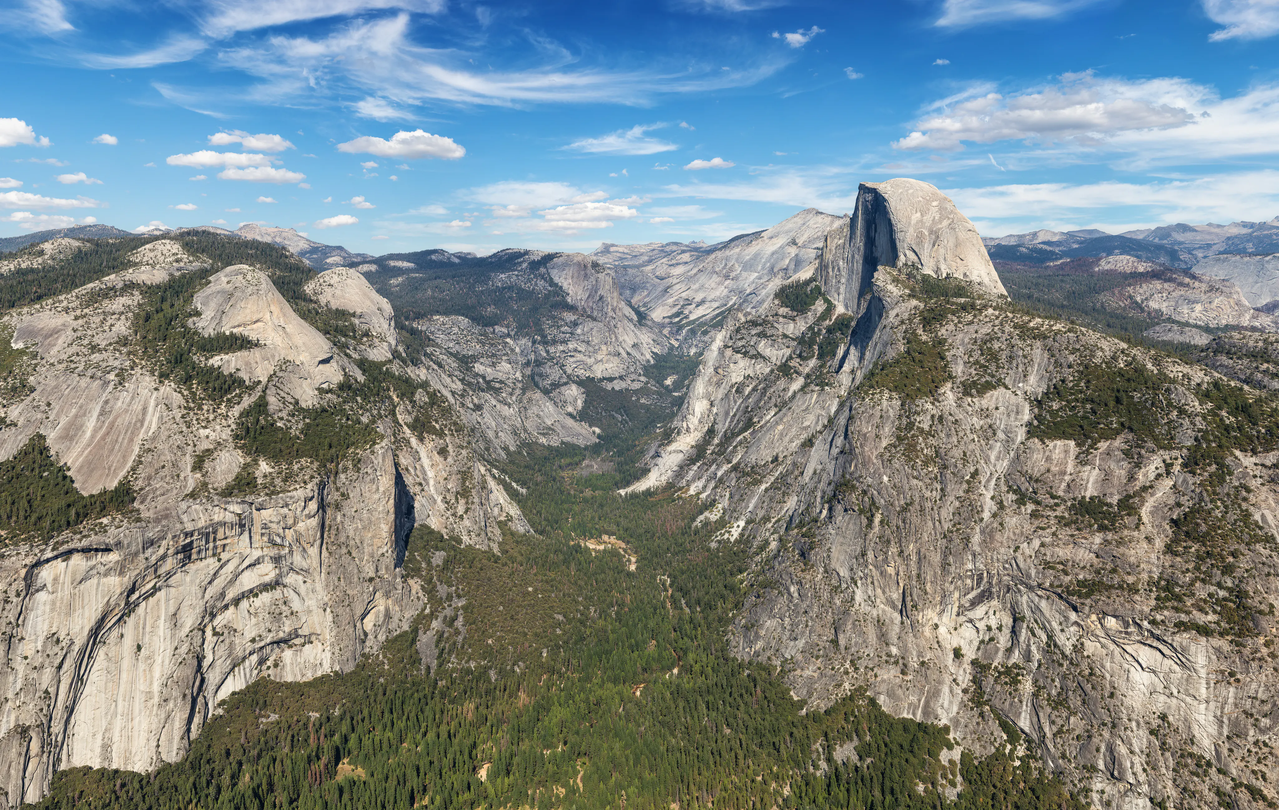 A panoramic view of Half Dome and Eastern Yosemite Valley with granite mountains, green forests, and a clear blue sky with scattered clouds