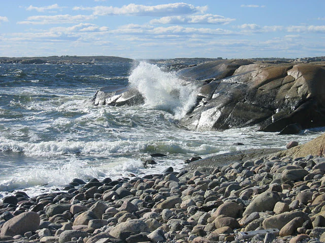 Ocean waves crashing against rocky shore with pebbly beach under partly cloudy sky