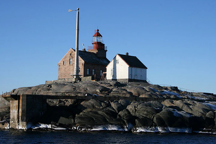 A red-topped lighthouse with adjacent buildings sits on a rocky island surrounded by water under a clear blue sky