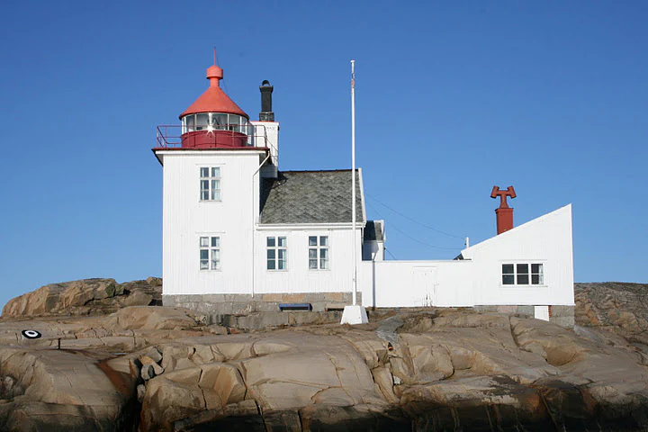 White lighthouse with red roof on rocky terrain under clear blue sky