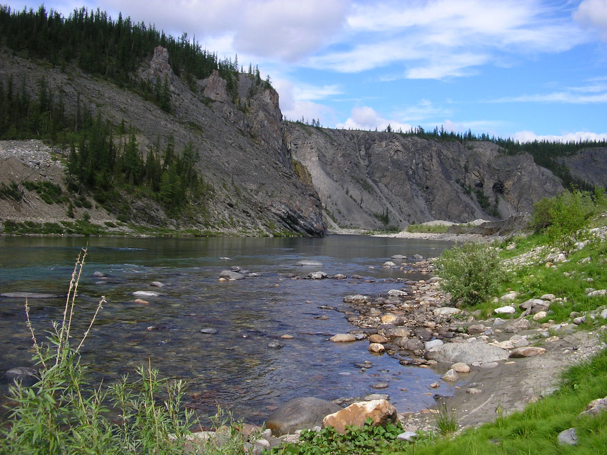 A wide shot of a river flowing through a valley with a rocky cliff face covered in sparse evergreen trees on the left side.