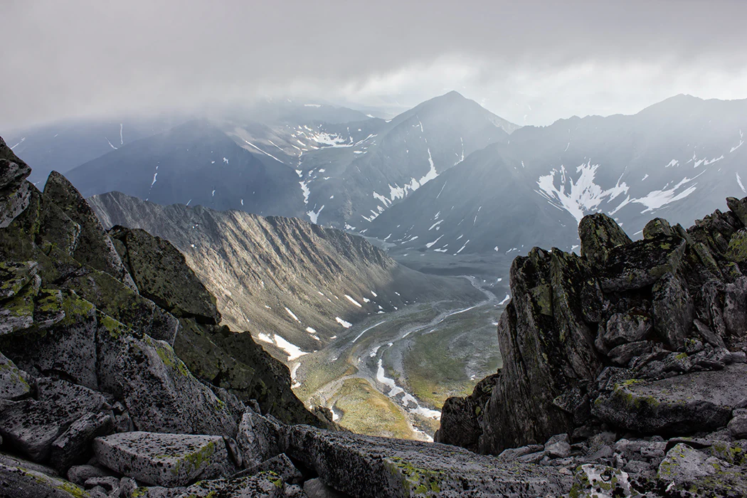 A wide, high-angle view looking down from rocky, gray mountain slopes into a deep valley with patches of snow and a river, under an overcast sky.