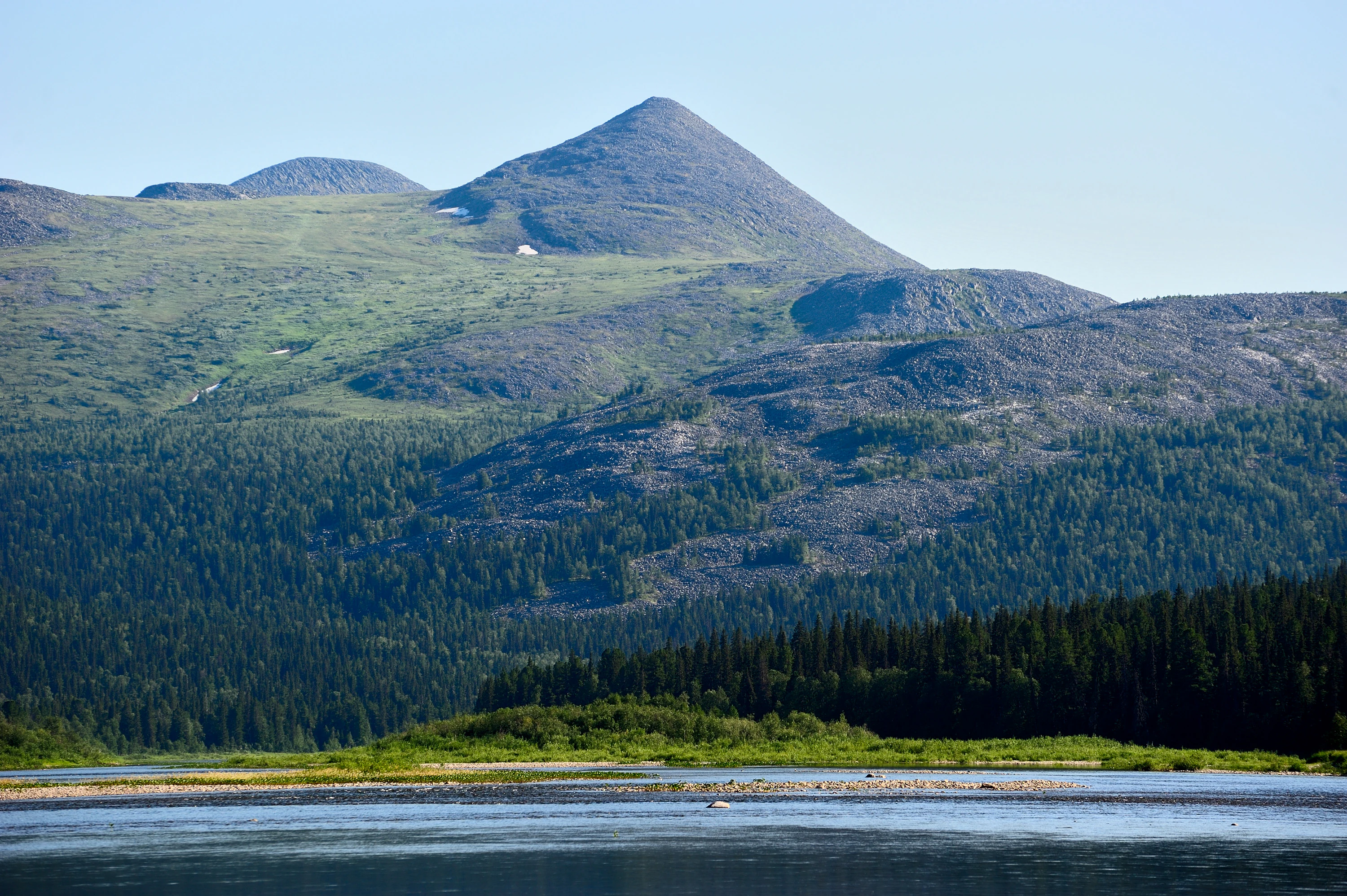 A wide scenic view of the Shchugor River in the foreground with a coniferous forest and the rounded, green slopes of the Uuty Ridge rising in the background under a clear sky.