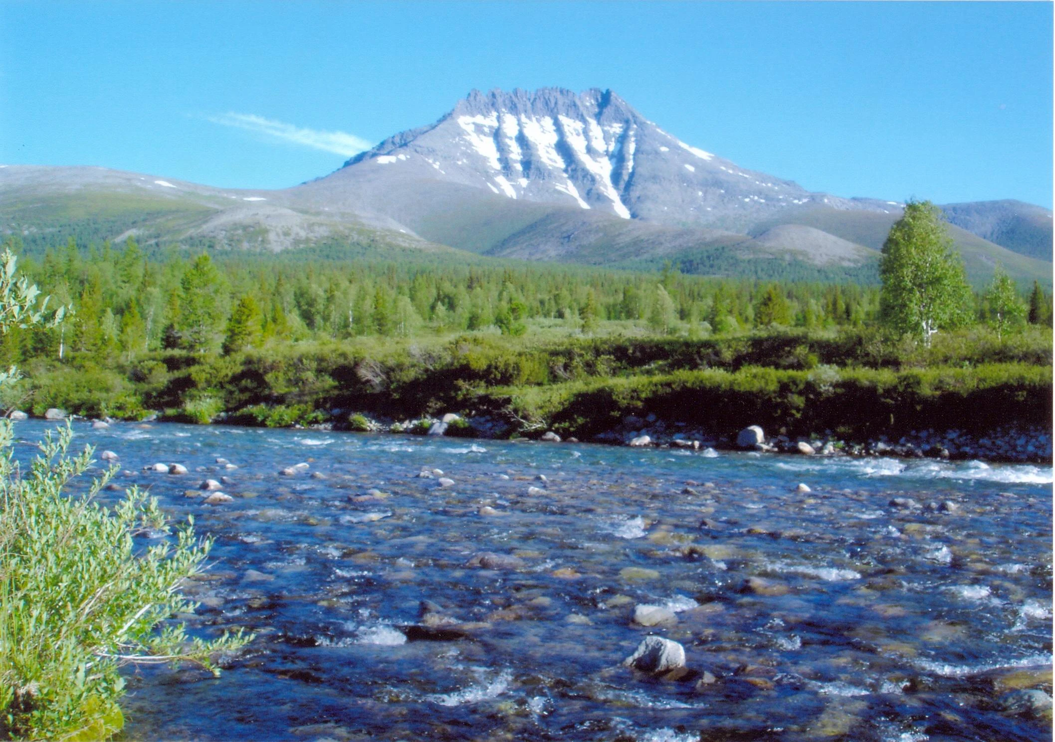A wide shot of a shallow river flowing over a rocky bed, with a grassy riverbank and a large, snow-streaked mountain peak in the background under a clear blue sky.