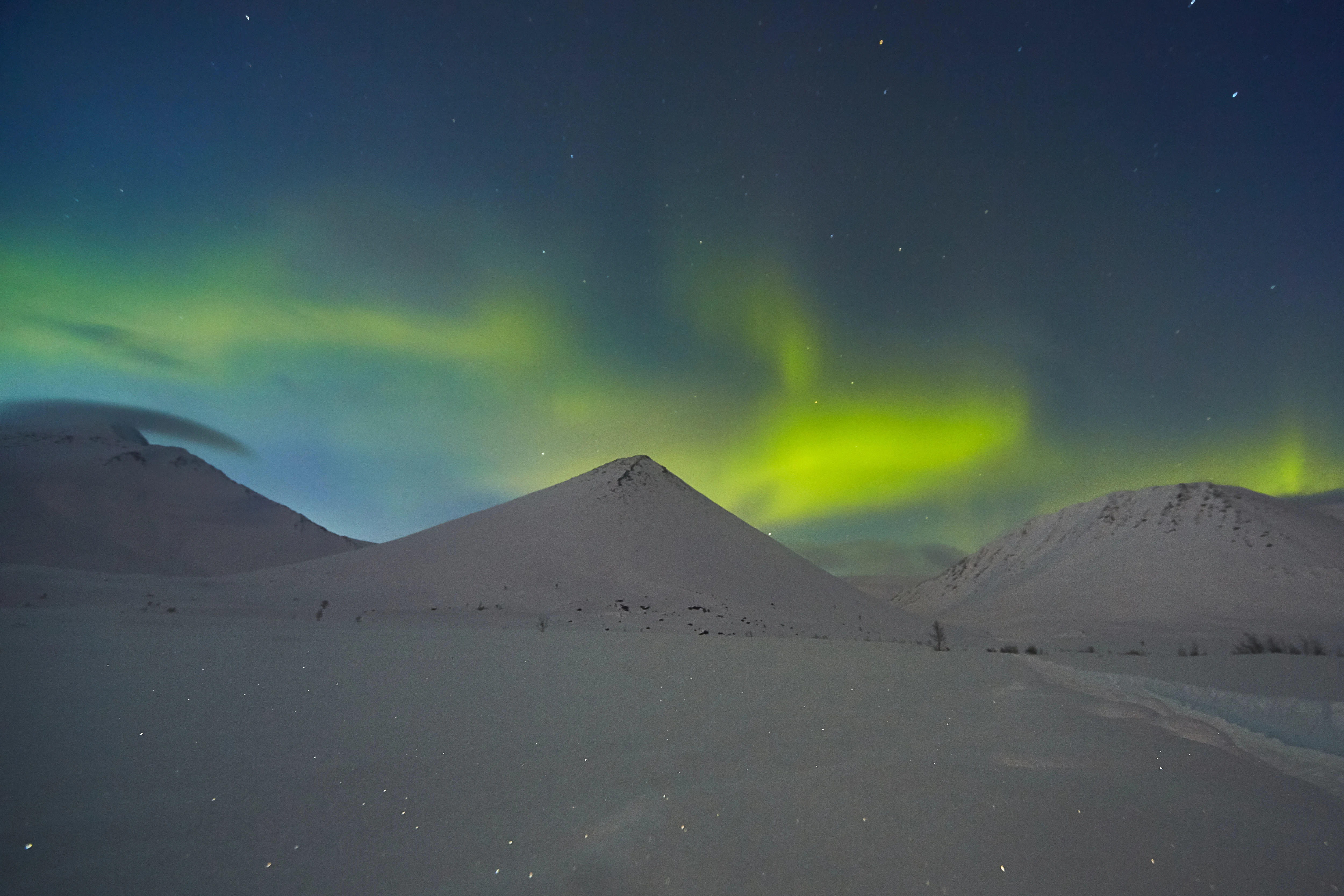 A night landscape showing bright green aurora borealis lights dancing above dark, snow-covered mountain ridges in a remote polar region.