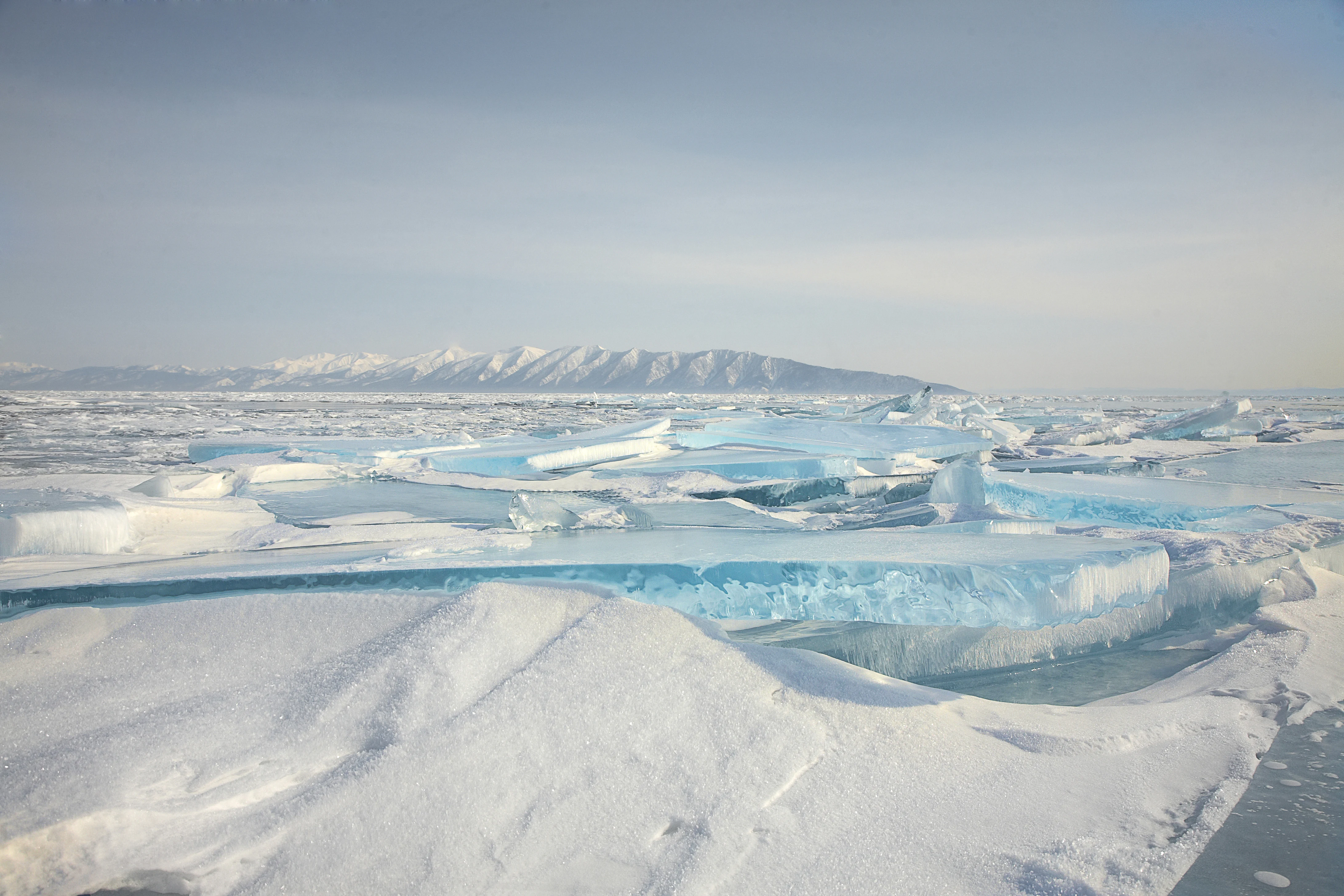 Extensa superficie de lago congelado con placas de hielo azul rotas y dentadas en primer plano, y una lejana cadena montañosa cubierta de nieve bajo un pálido cielo invernal.