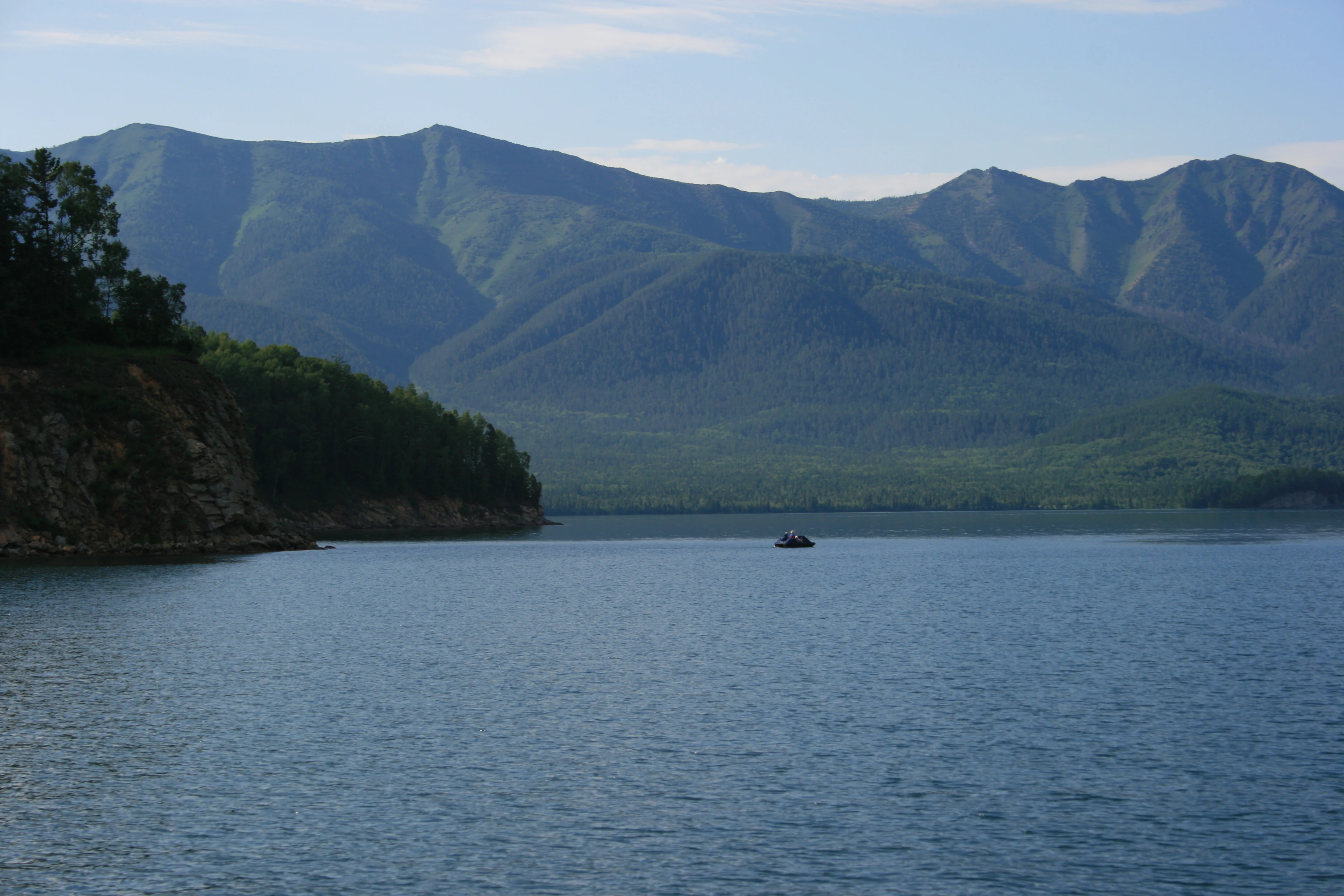 Calm blue lake water with a forested shoreline and mountain range under a clear sky