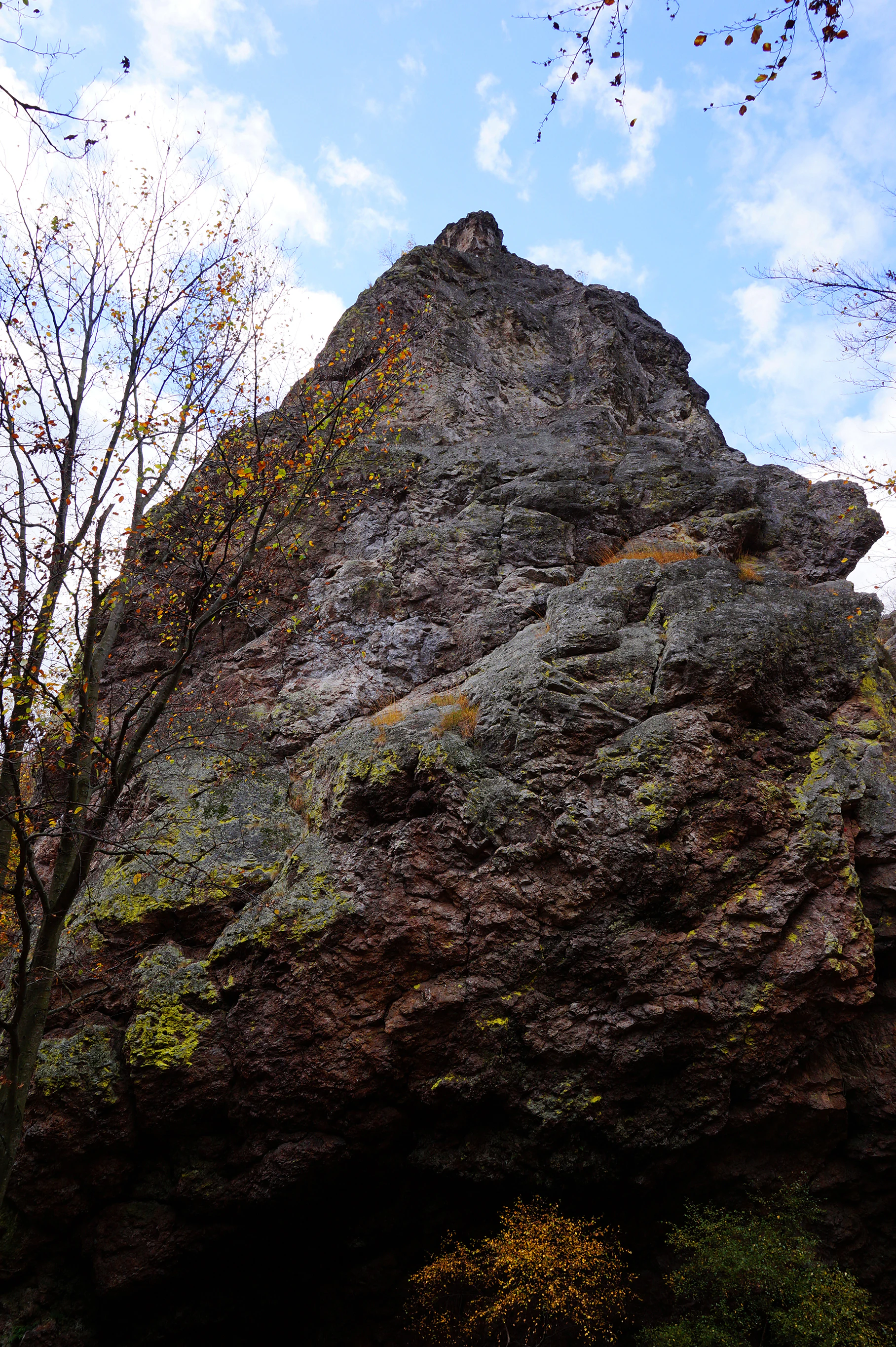 Large rocky mountain formation with moss and lichen, surrounded by trees with autumn foliage, under a partly cloudy sky