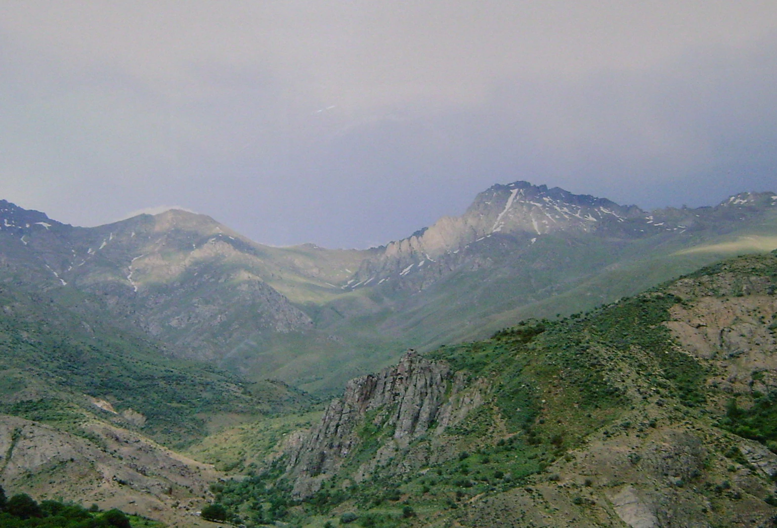 Panoramic landscape of mountainous terrain with rocky cliffs, green valleys, and distant peaks under an overcast sky