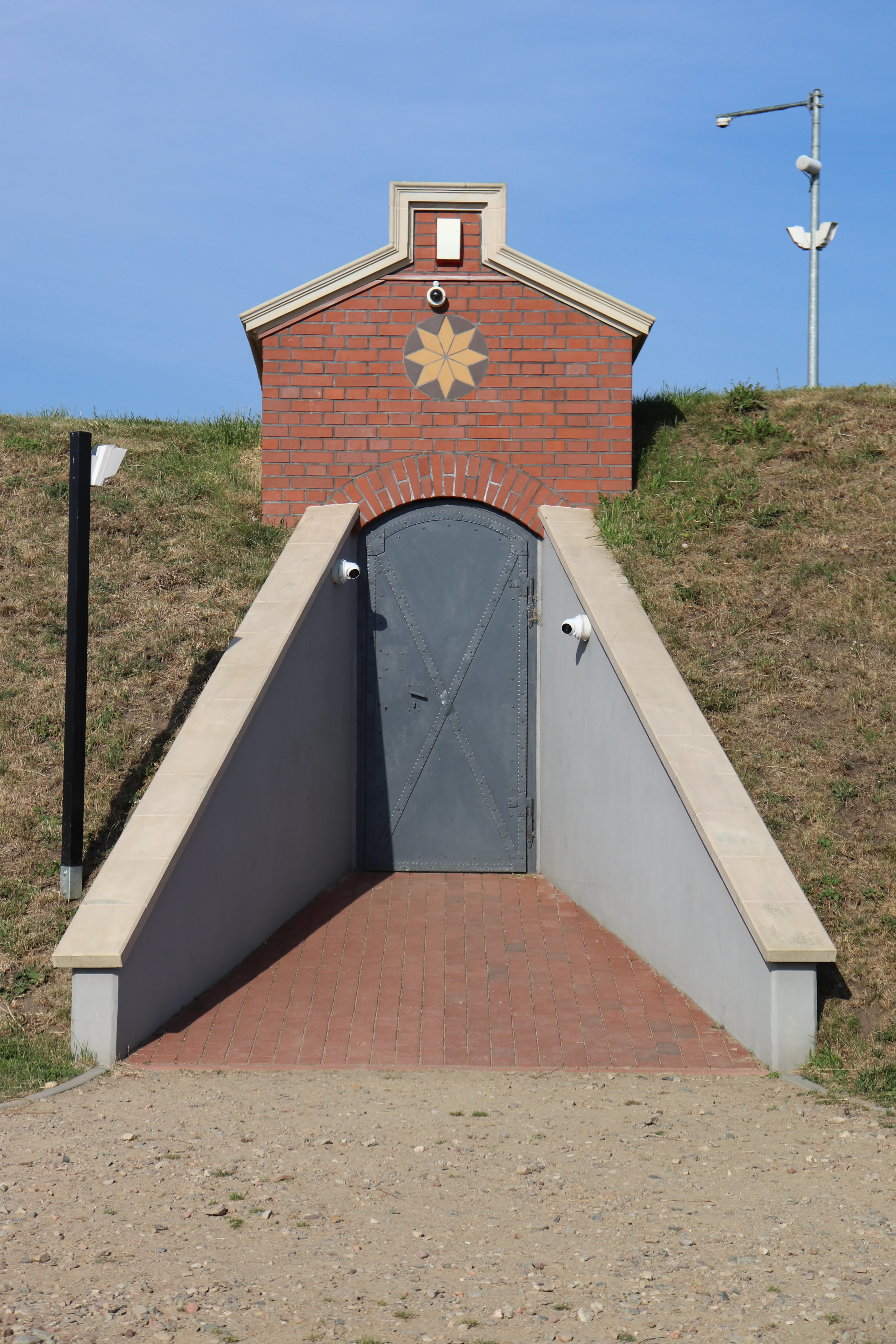 A red brick building entrance built into a grassy hill, featuring a gray arched metal door and a star-shaped ornament above the entrance.