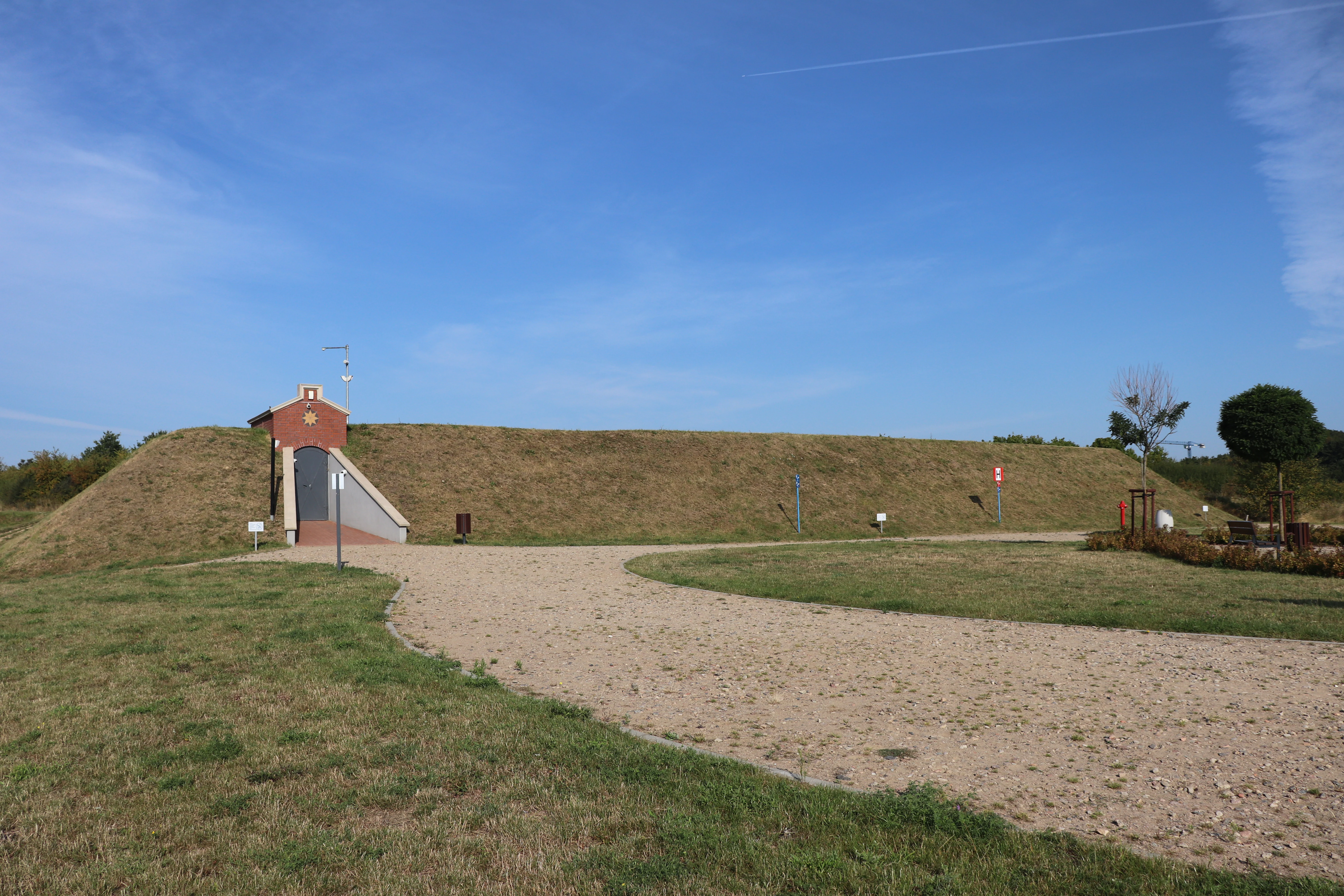 A red brick building entrance built into a grassy mound, serving as an access point for the Stara Orunia water reservoir, with a gravel path leading up to it under a clear blue sky.