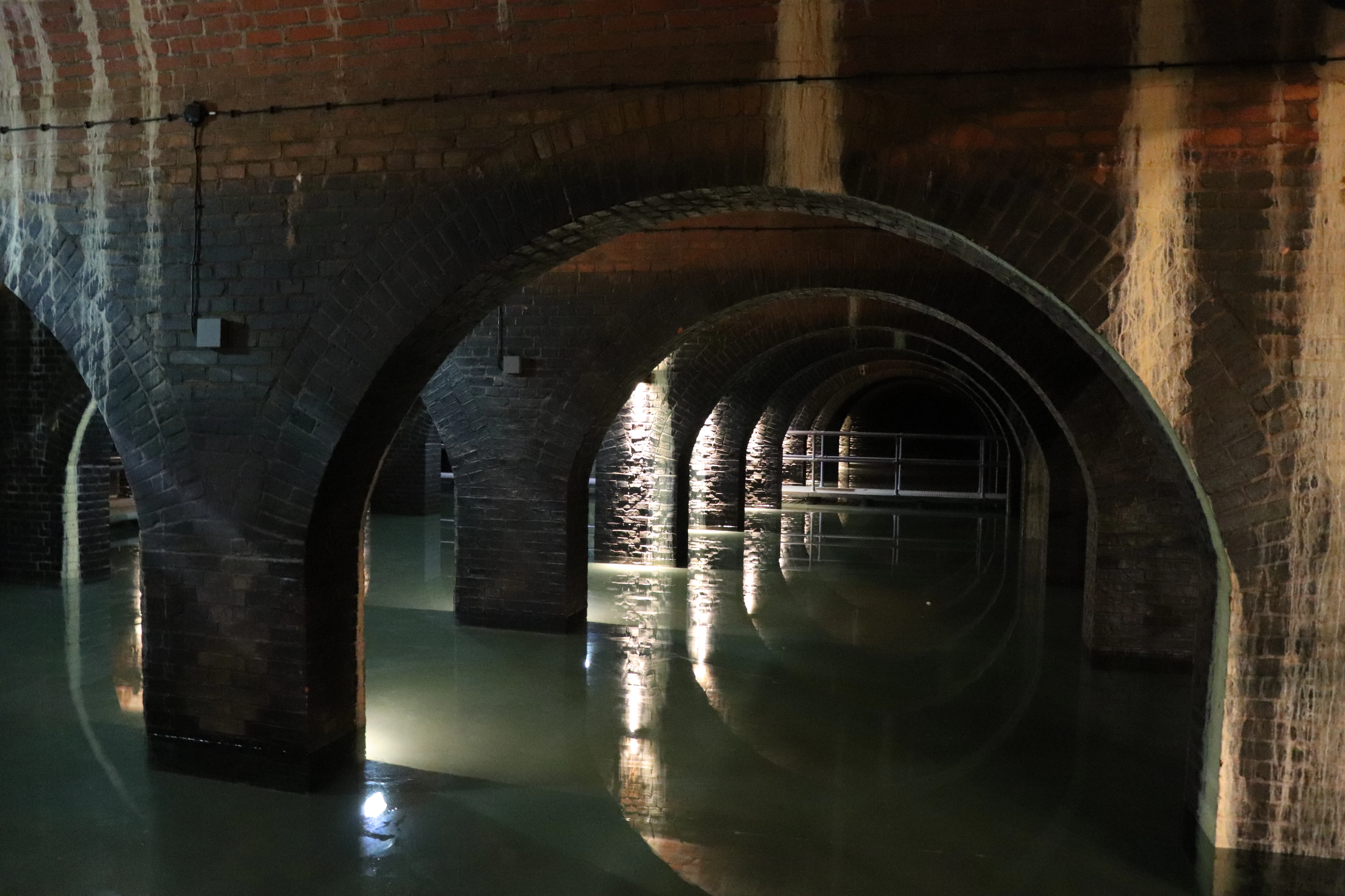A wide, low-angle shot inside the Stara Orunia water reservoir, showing multiple rows of dark, red-brick arches reflecting in a still pool of water, illuminated by warm artificial lights.