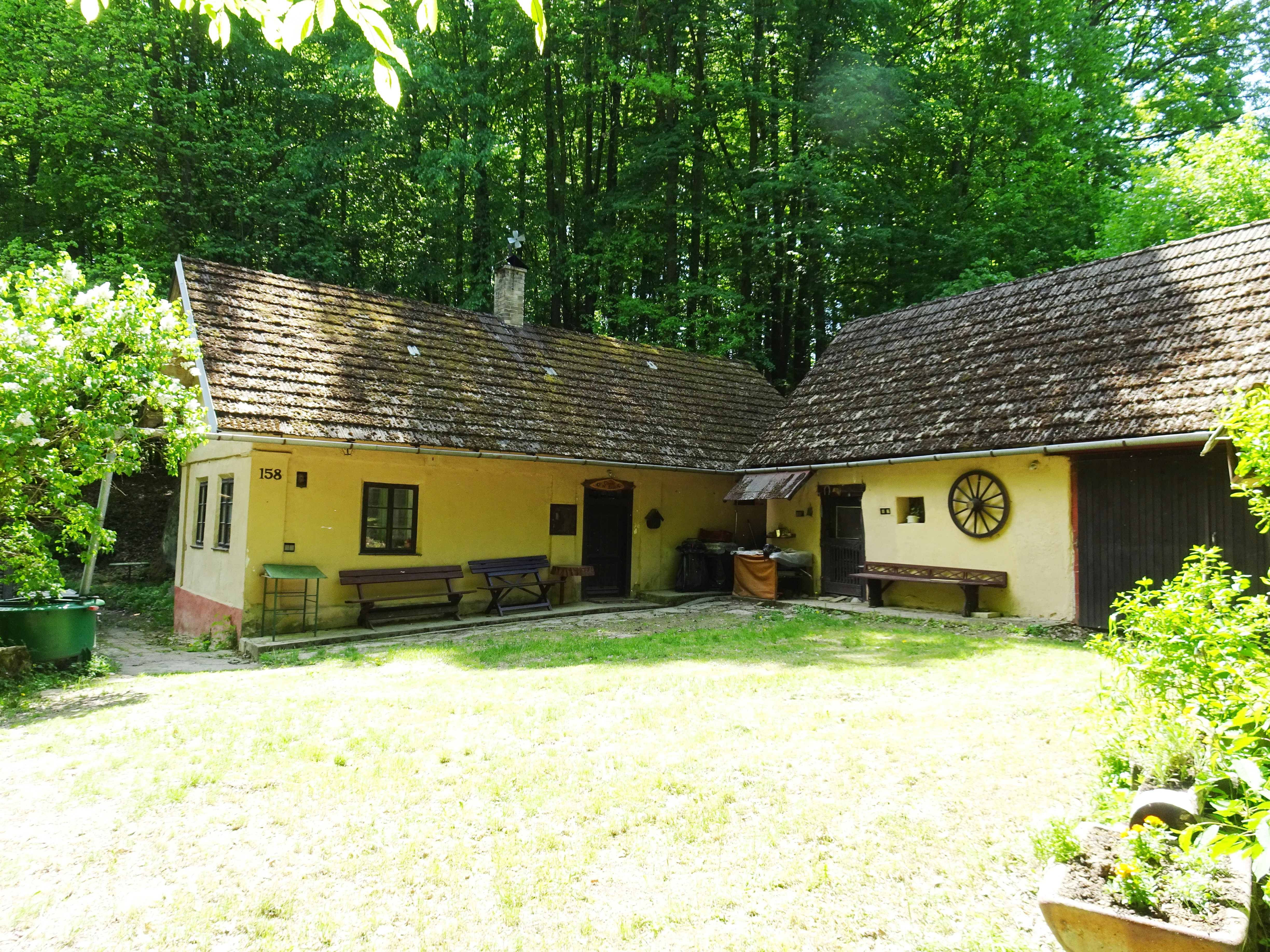 A rustic yellow historic cottage with a brown tiled roof and wooden wagon wheel decoration, situated in a green, wooded setting.