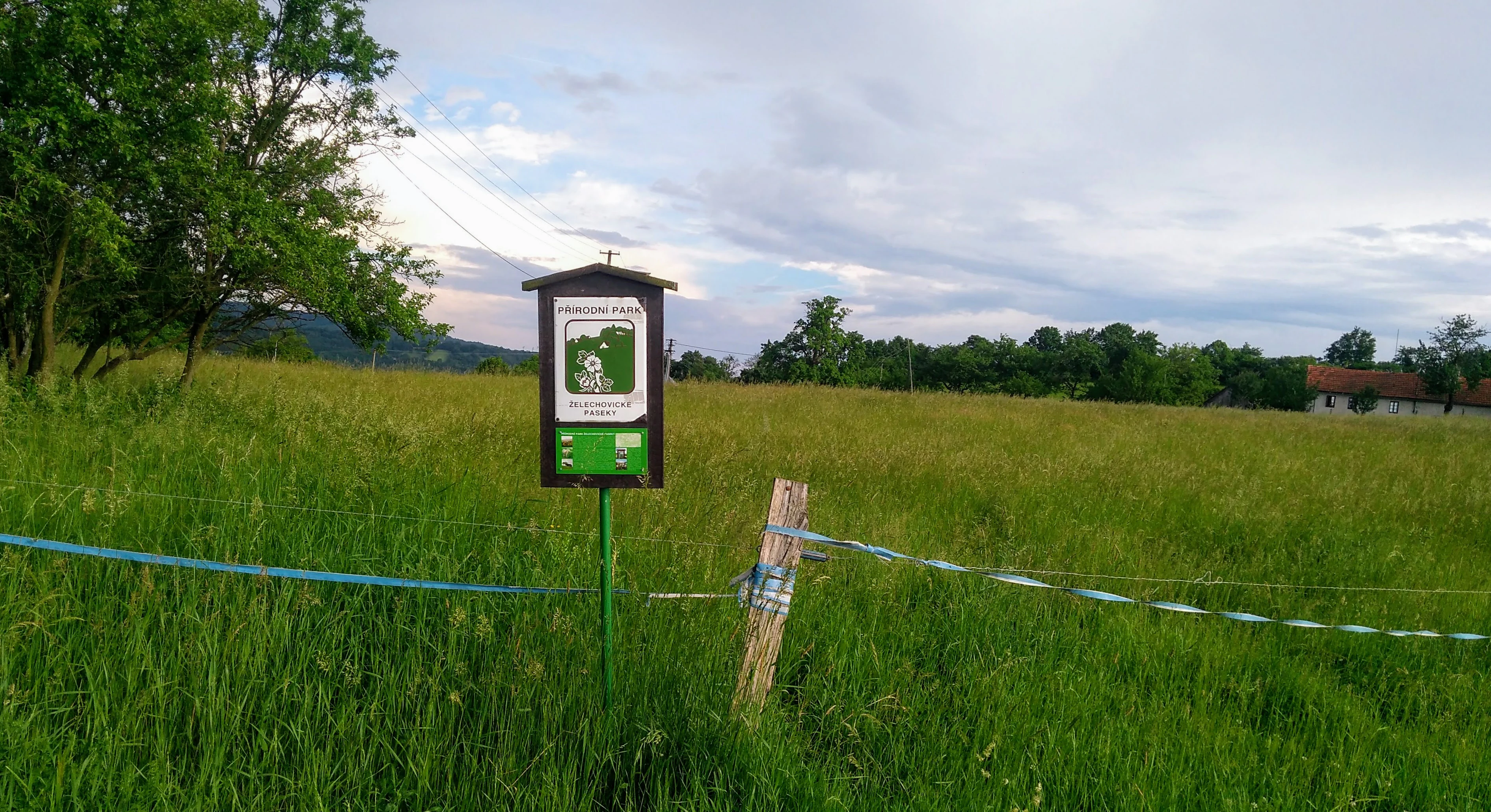 A green and white sign marking the nature park stands in a lush green meadow, with a makeshift fence and trees in the background under a cloudy sky.