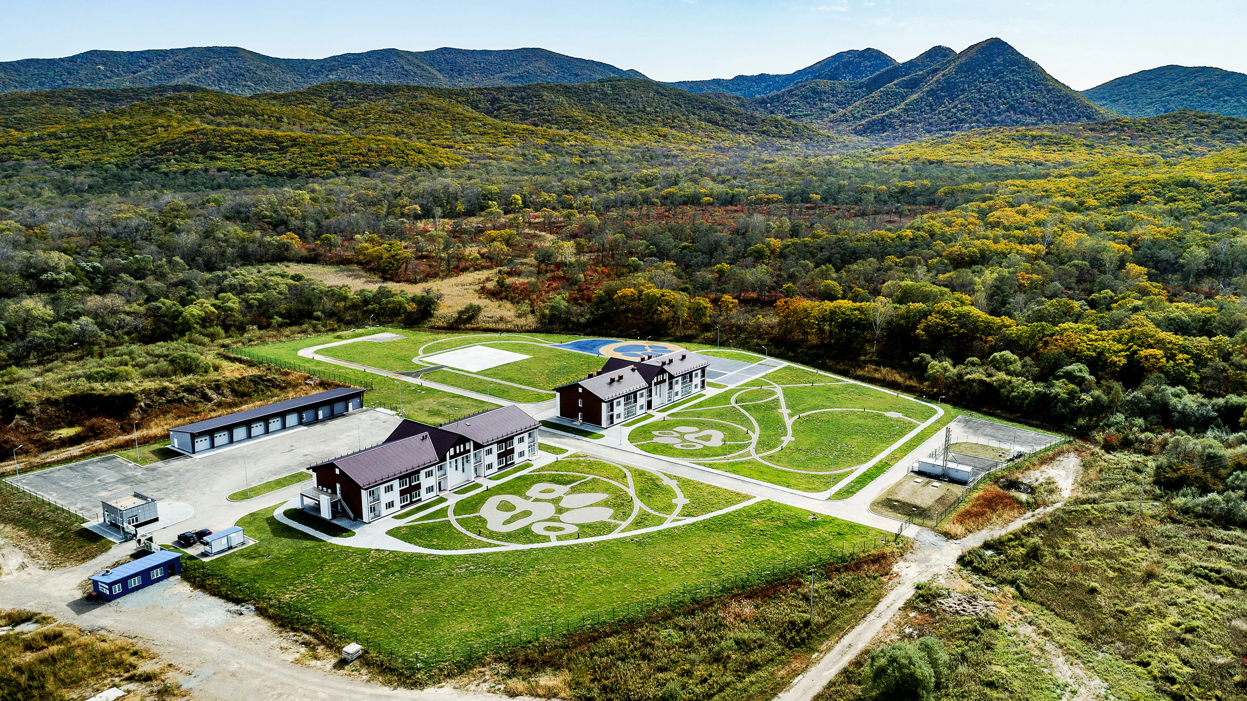 An aerial photograph showing a complex of buildings with dark roofs surrounded by green lawns featuring large white floral designs, set against a backdrop of forested mountains.
