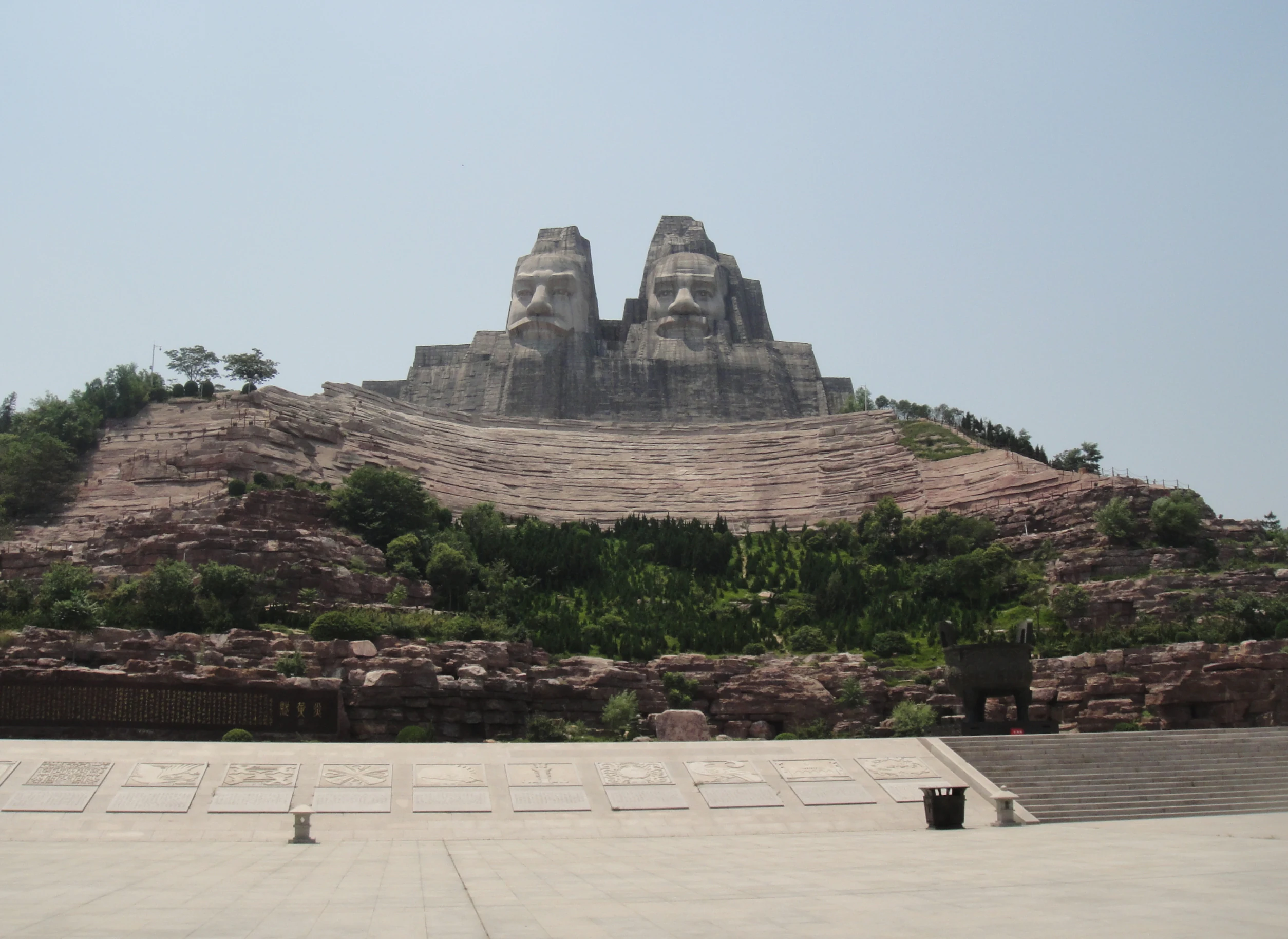 A wide angle shot of the massive stone sculptures depicting two large human faces carved into the top of a hill, overlooking a tiered plaza at the Yellow River Scenic Area.