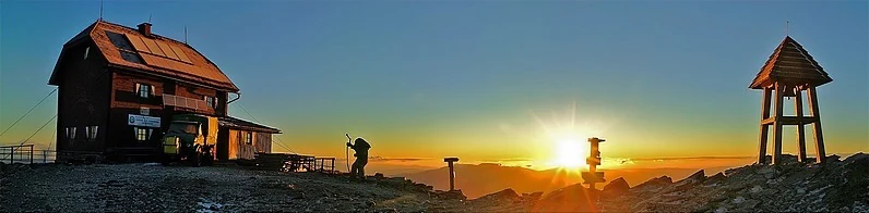 Panoramic view of a mountain summit at sunrise, featuring a wooden hut on the left, a hiker in silhouette, and a small wooden bell tower on the right, with a bright sun rising over distant peaks.