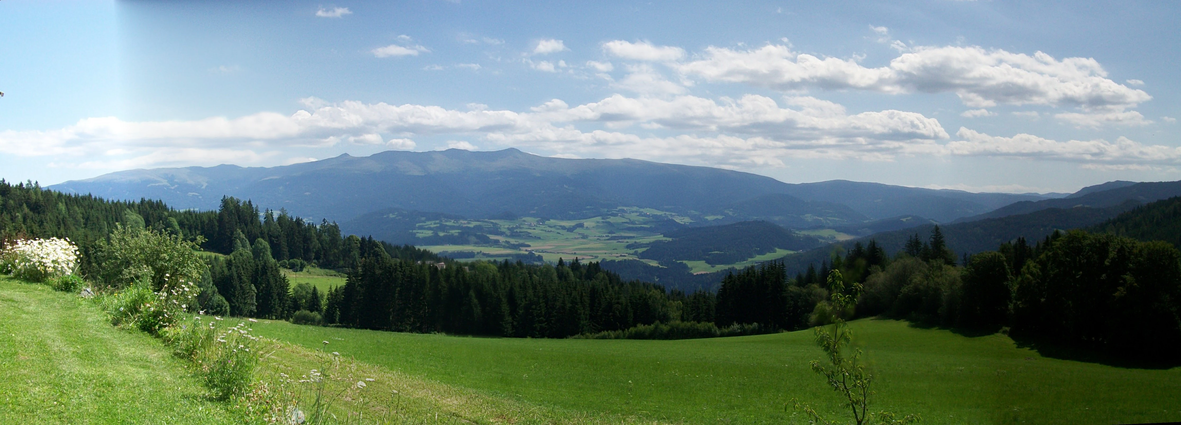 Panoramic landscape view of the Zirbitzkogel mountain peak rising above green rolling hills and evergreen forests under a blue, partly cloudy sky.