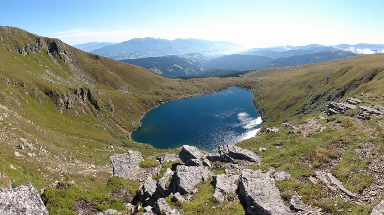 A high-angle view looking down into a mountain cirque containing a deep blue lake, surrounded by grassy slopes and rocky outcrops.