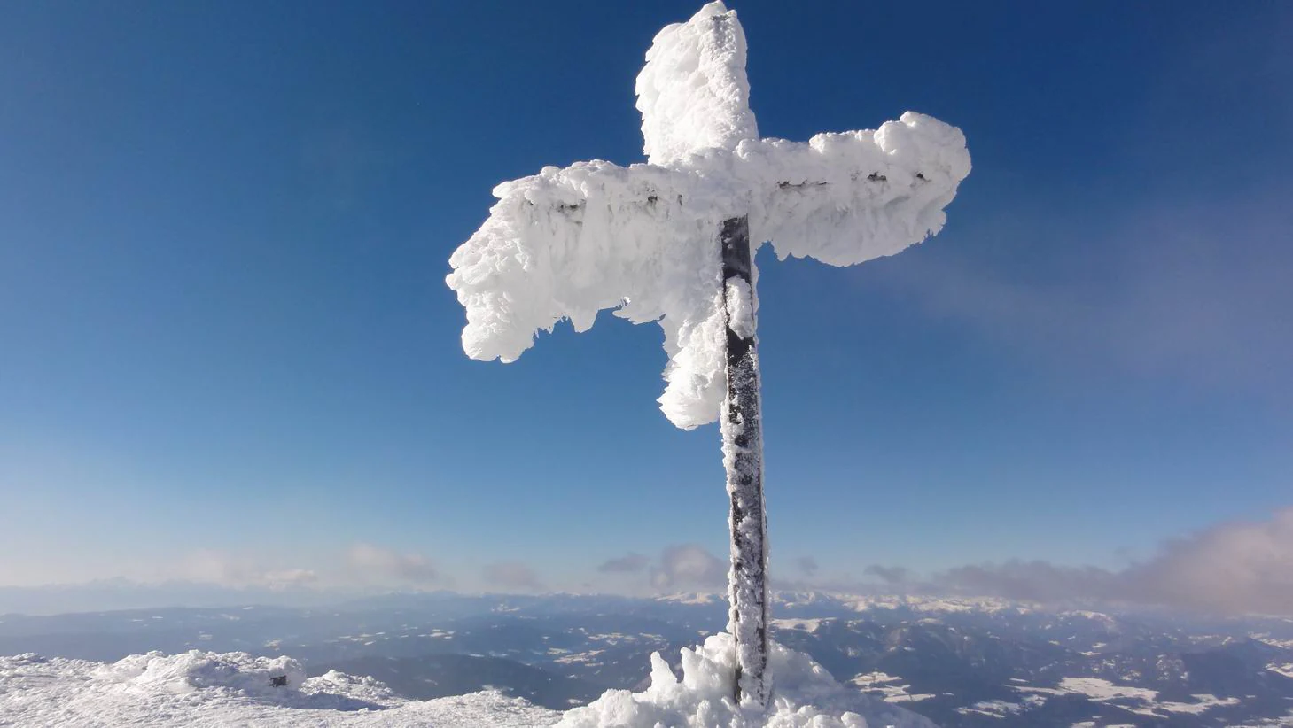 A thick, rime-covered mountain summit cross stands against a clear blue sky, overlooking a snowy mountain landscape.