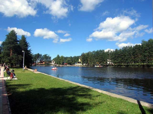 Grassy shoreline, lake, and trees with a red boat on the water under a blue sky with white clouds