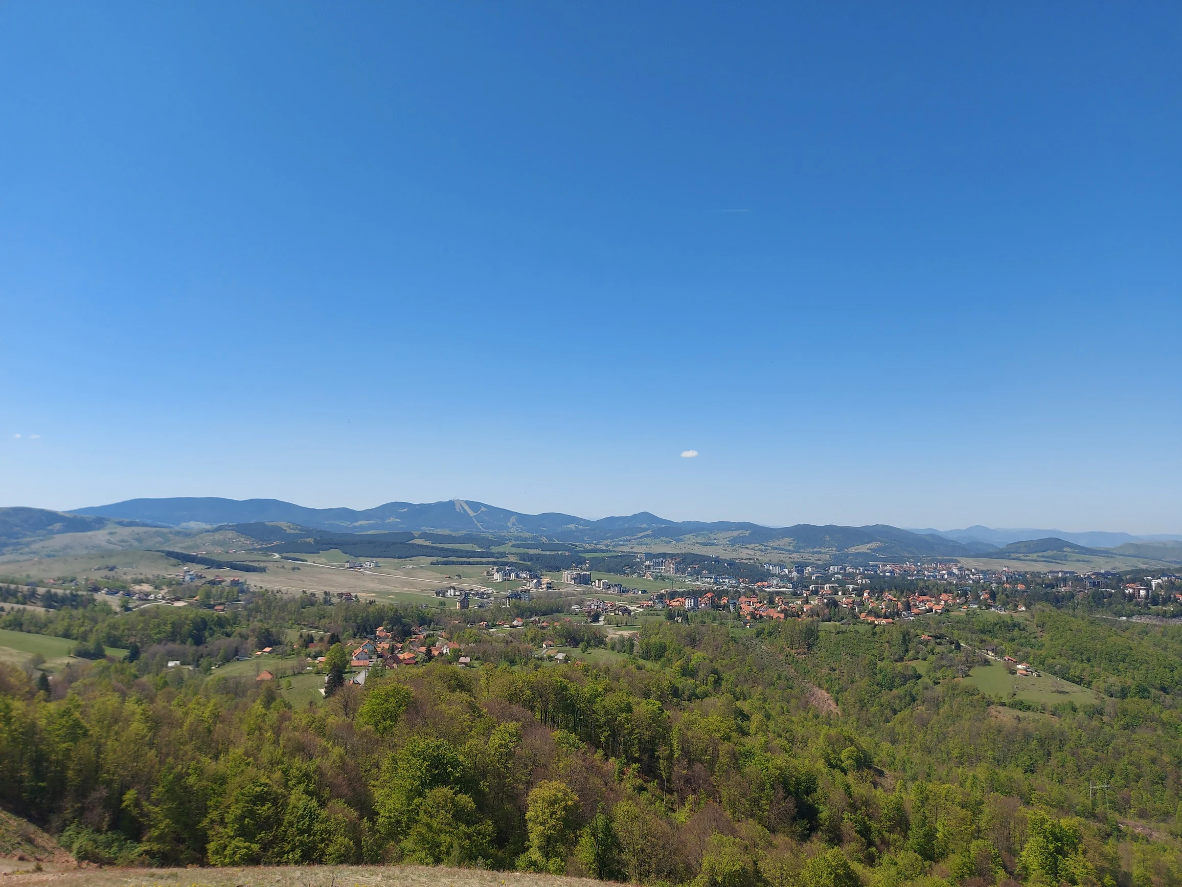 Wide landscape showing valley with town, green hills, and distant mountains under clear blue sky